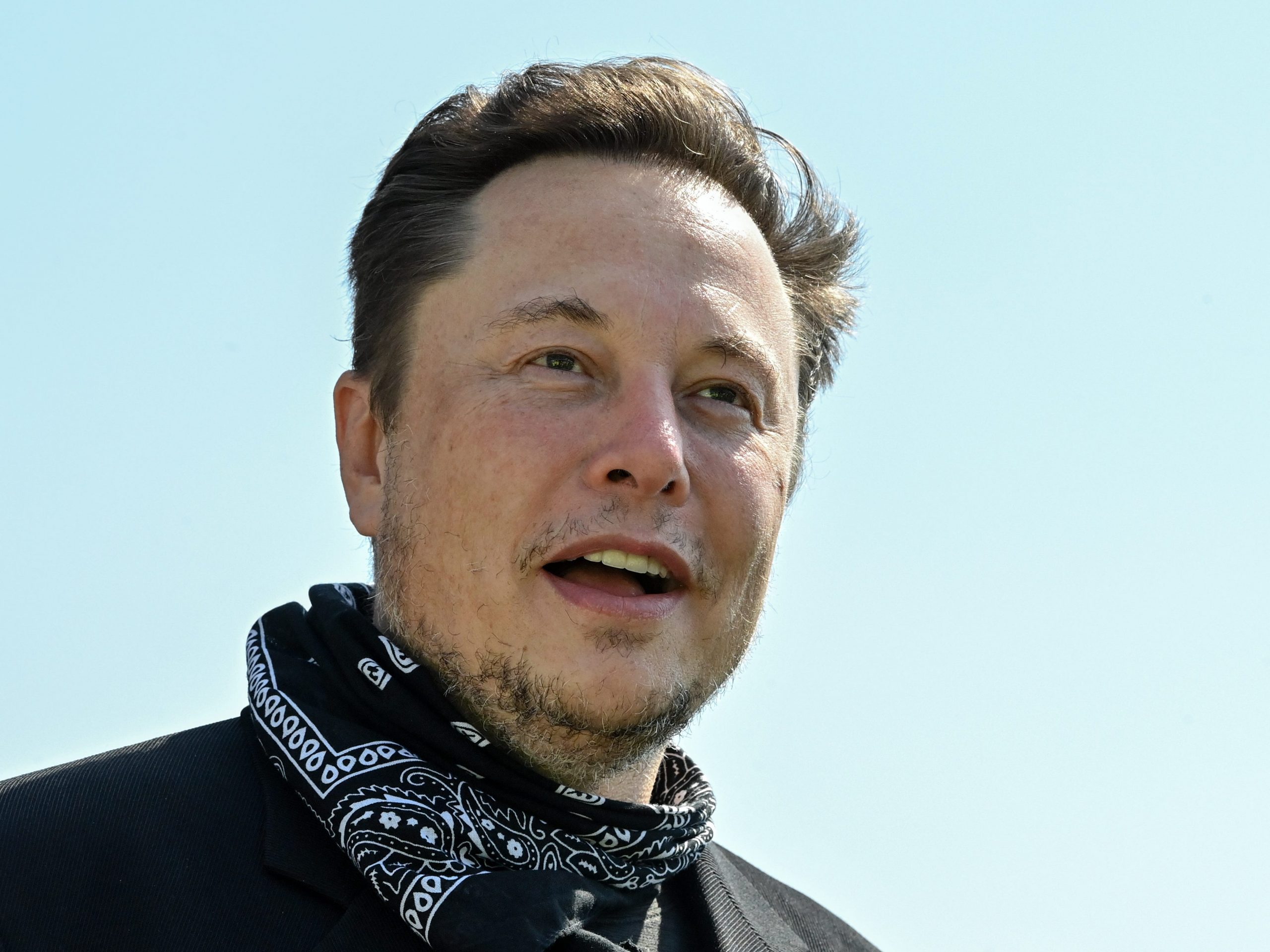 Tesla CEO Elon Musk wears a black and white bandana around his neck in front a light blue sky.