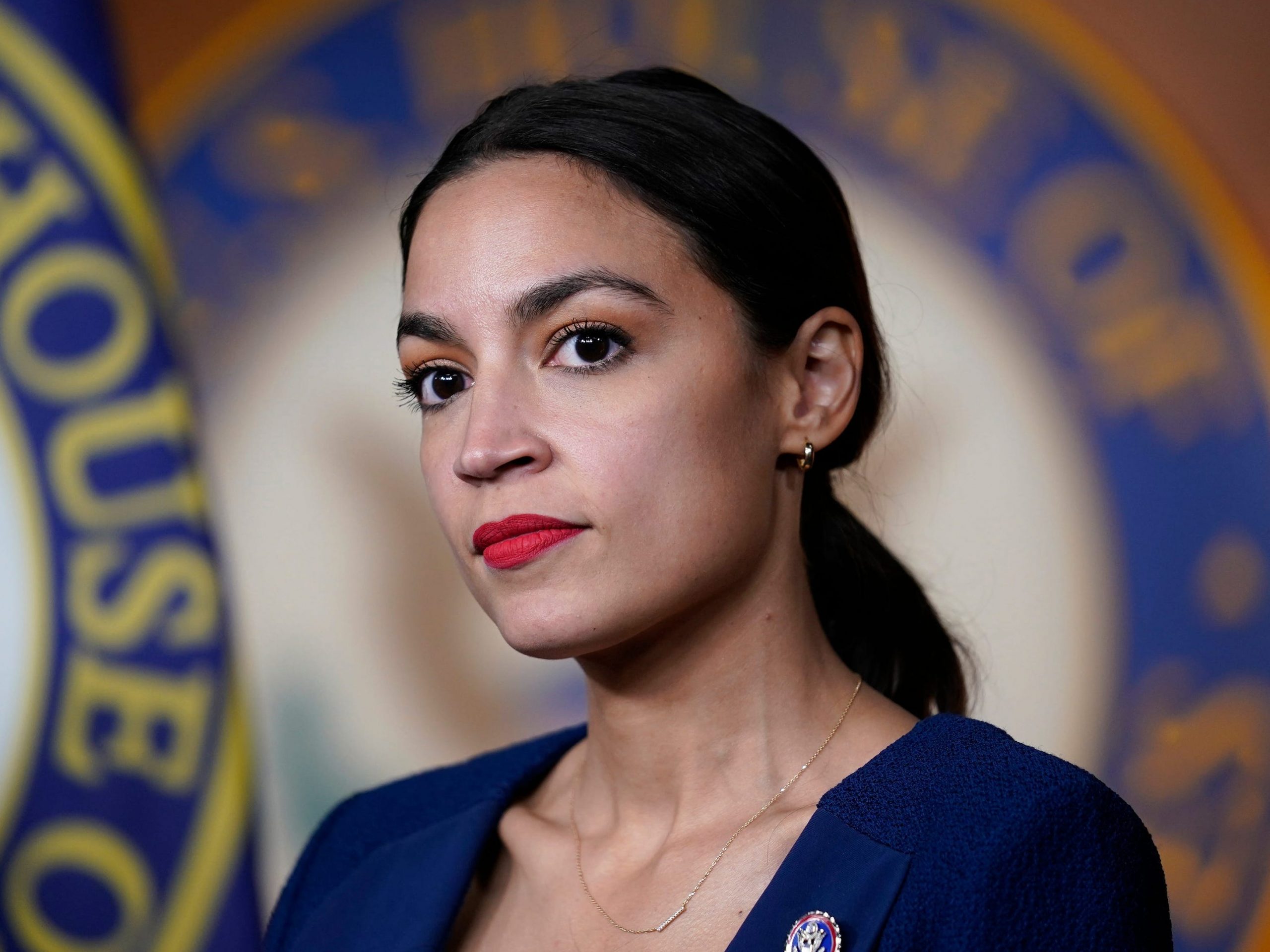 New York Congresswoman Alexandria Ocasio-Cortez in a blue blazer in front of a House of Representatives seal