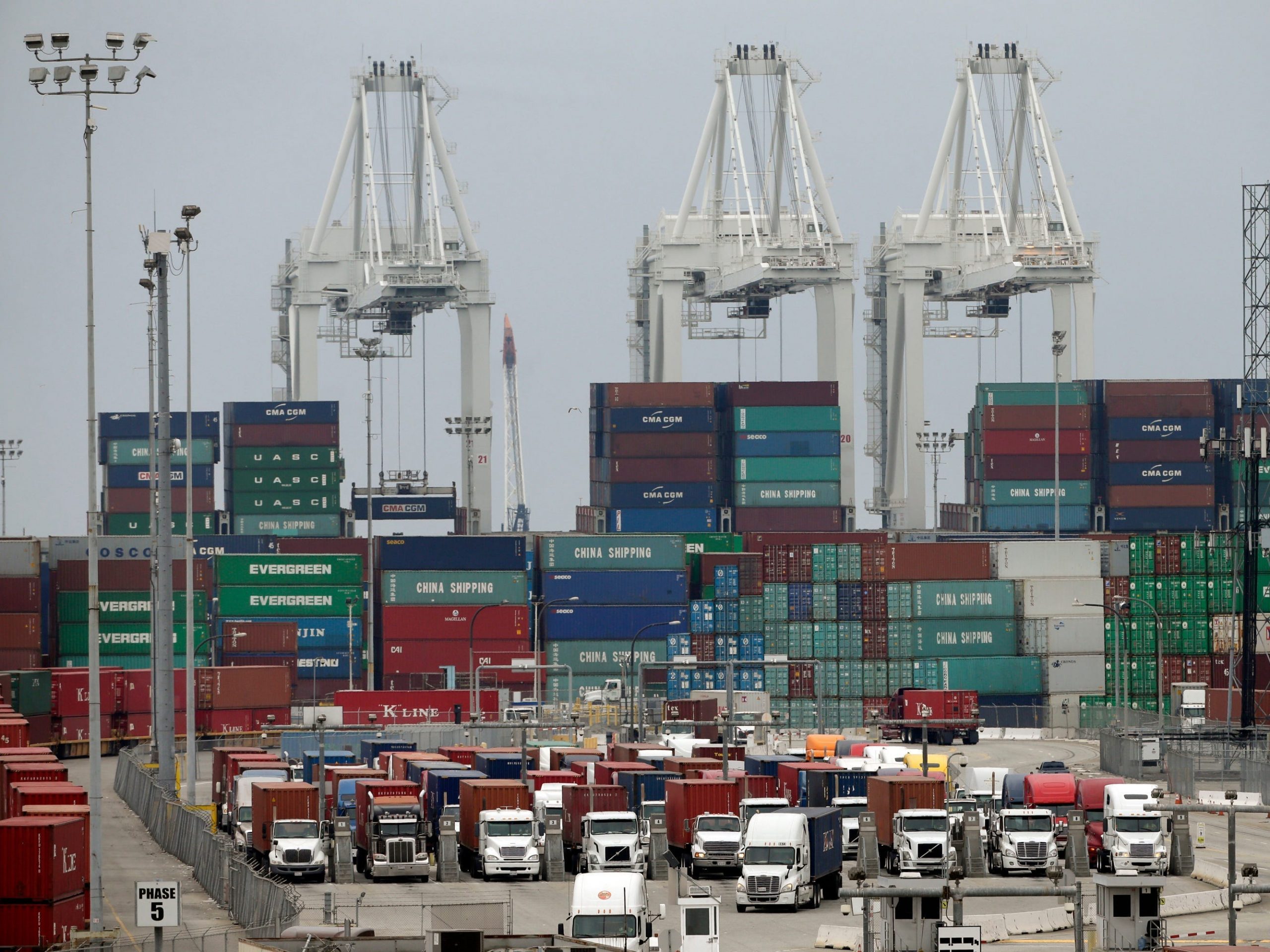 Several tractor trailers wait to depart a port with shipping containers stacked behind them