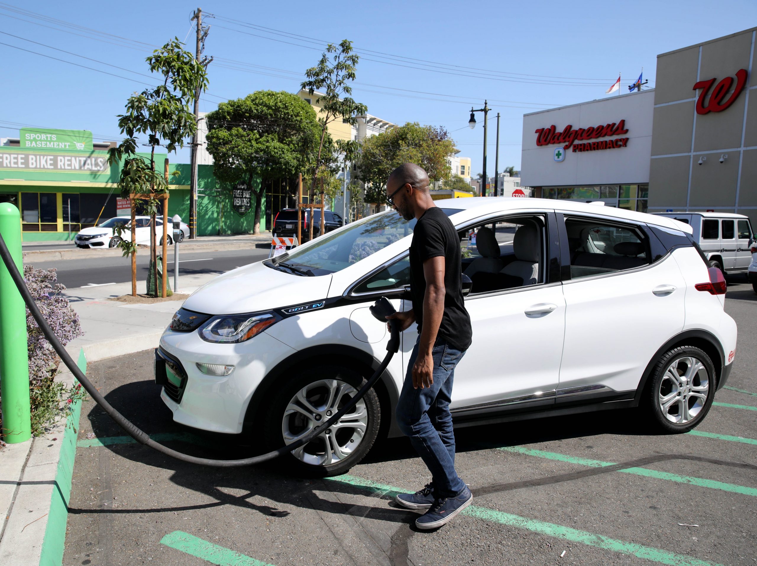 A man charges his white Chevrolet Bolt EV on a sunny day in a pharmacy parking lot.