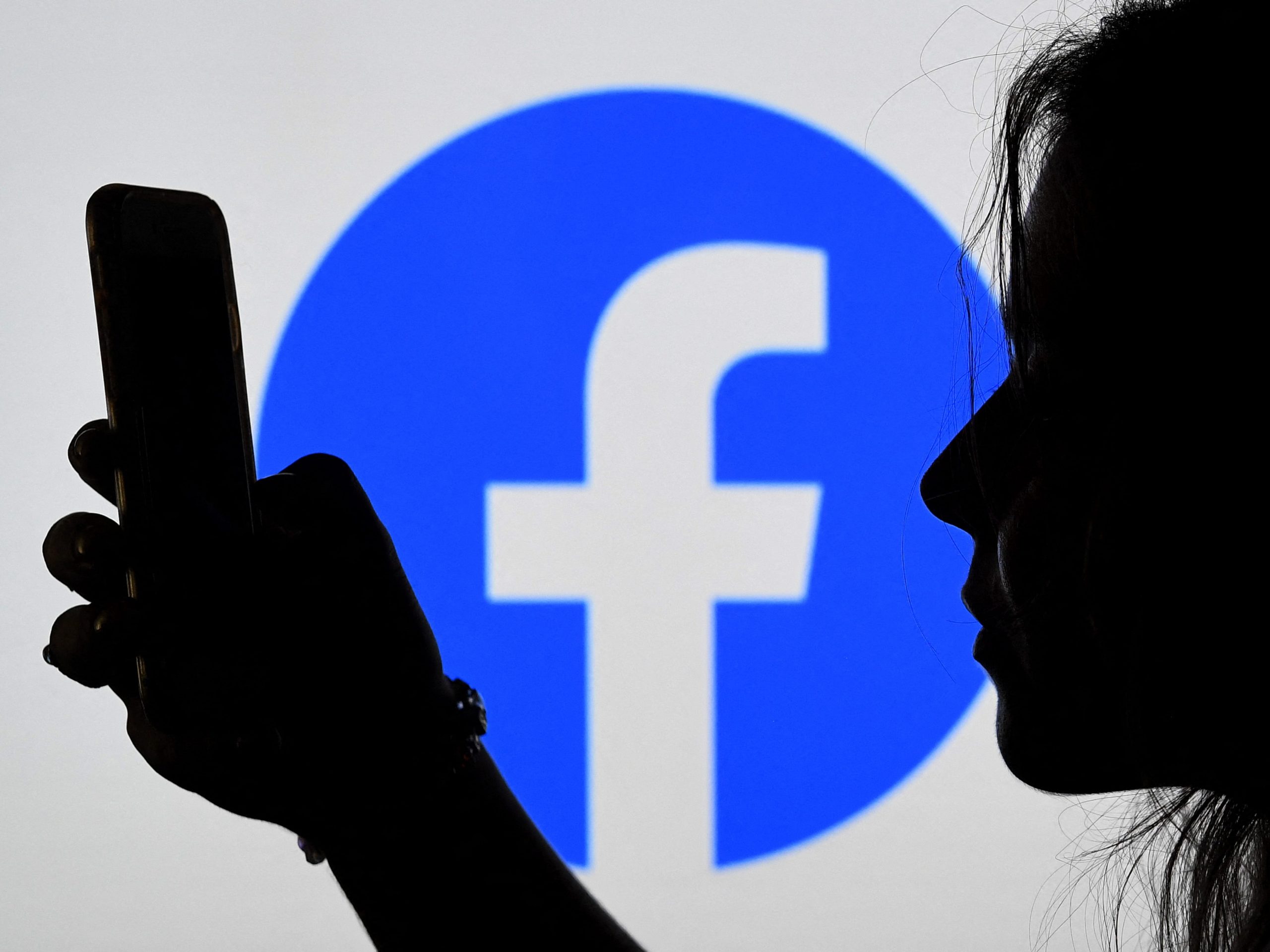 A person looks at. a smart phone with a Facebook logo displayed in the background