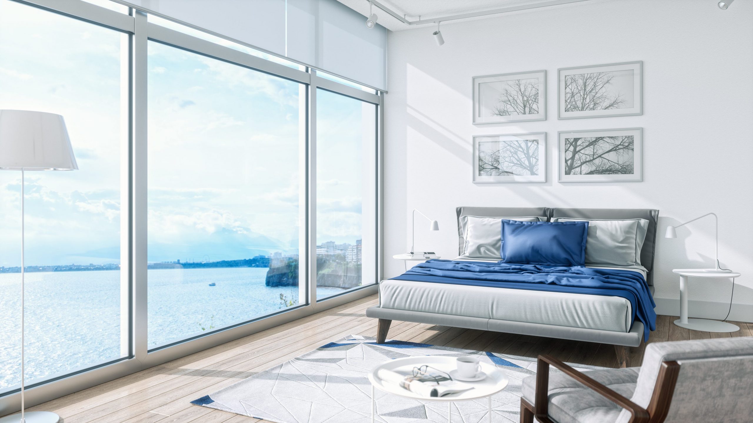 A bedroom with tall windows, a view of the ocean, and a bed dressed in gray and blue linen.