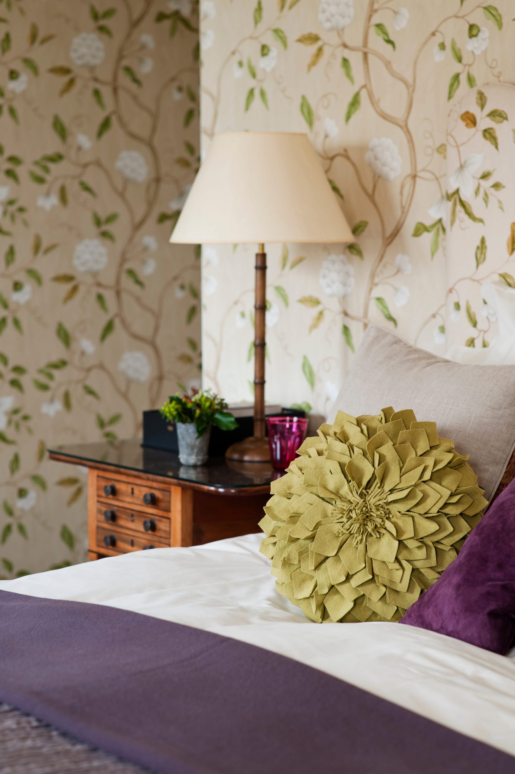 A close-up of a bed with purple linen and a gold pillow with a lamp and beige floral wallpaper in the background