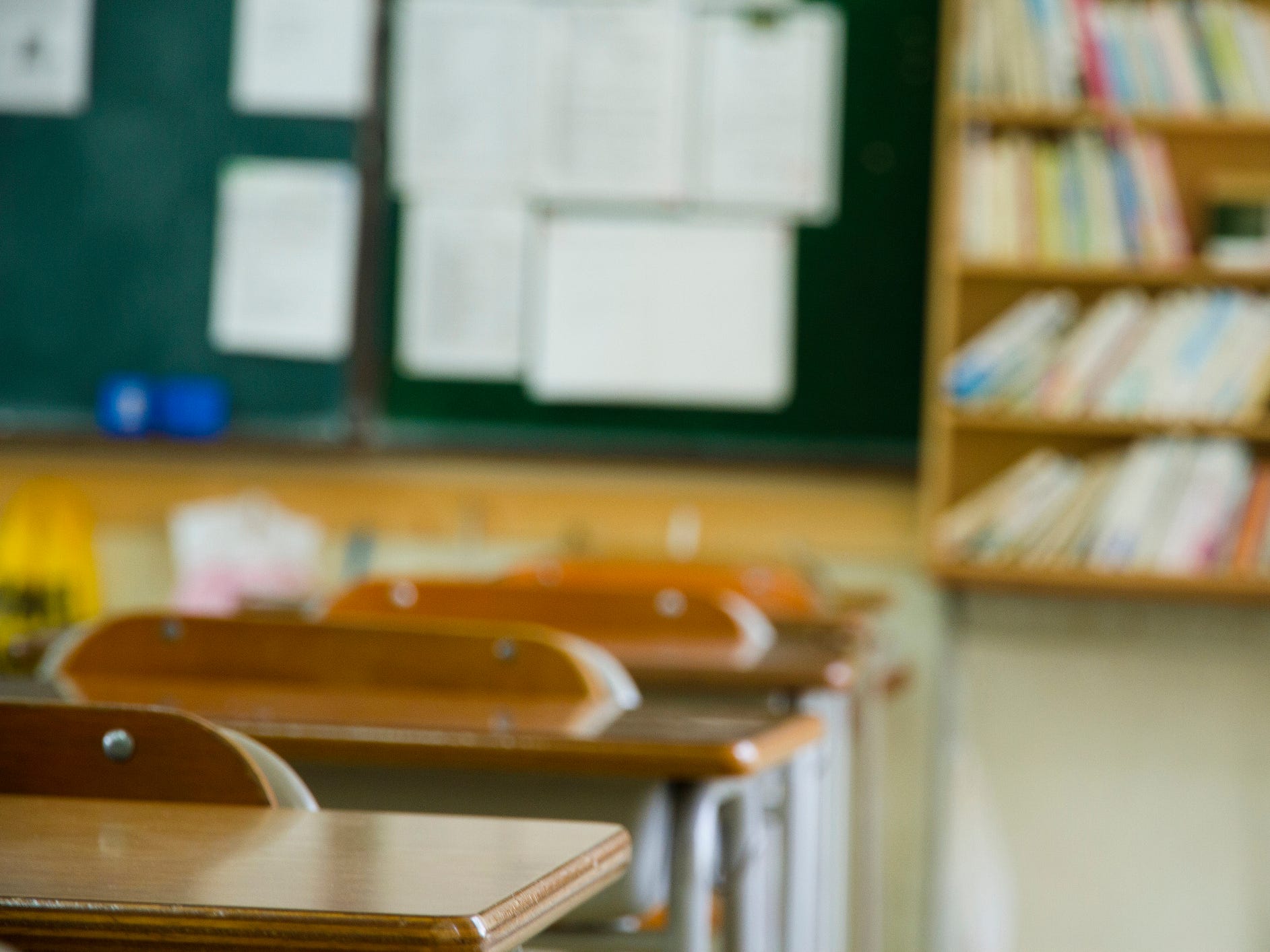 A school classroom, with desks, a blackboard, and bookshelves.