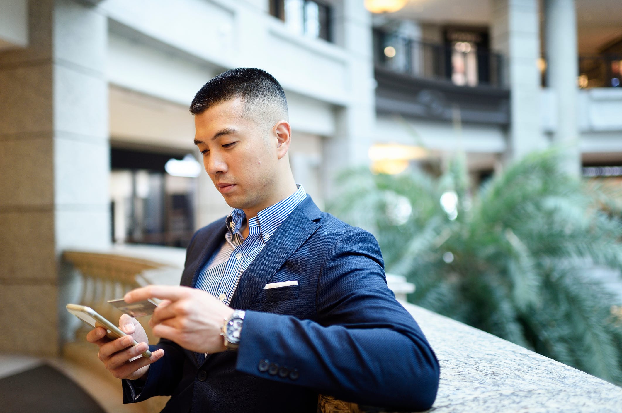 man sits in the lobby of a mall using his credit card to make a purchase on his phone