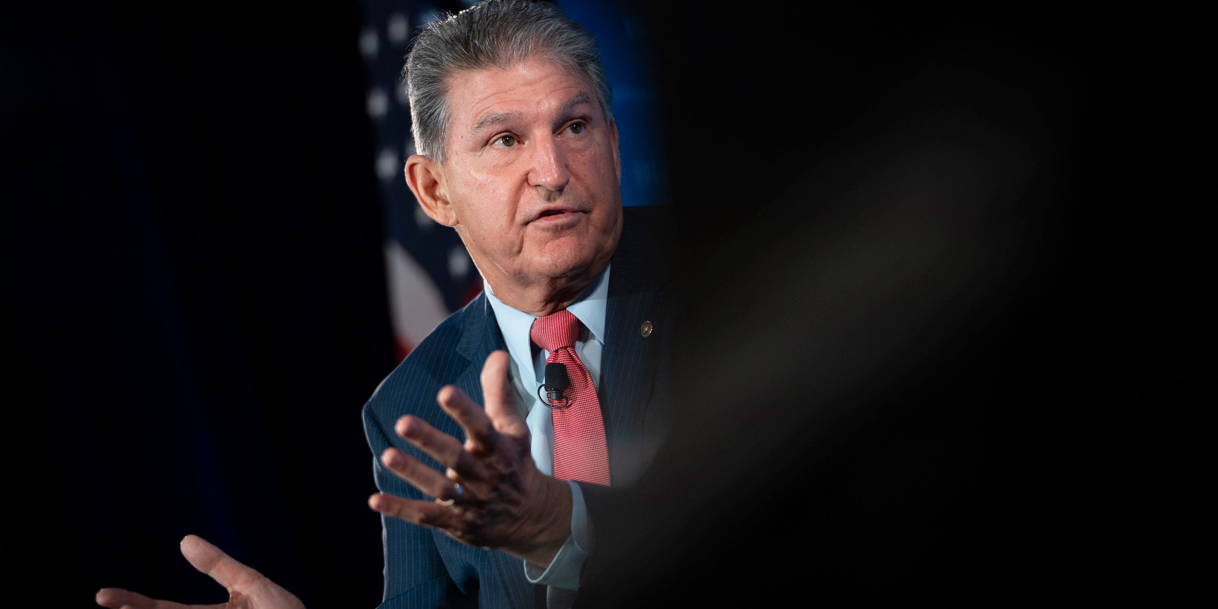 Senator Joe Manchin gestures in front of an American flag with his palms up, the right half of the photo is blurred out in black.