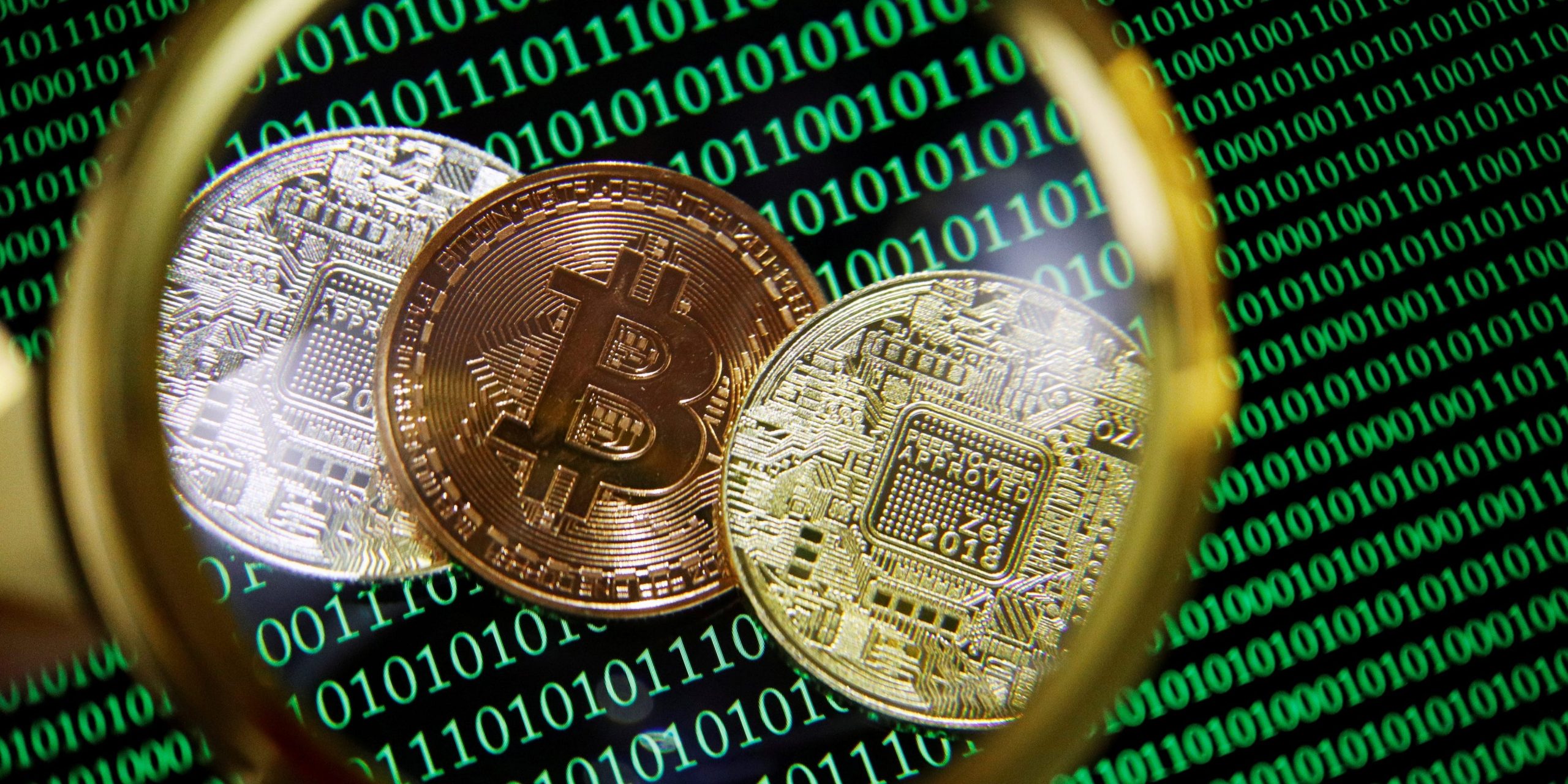 Representations of bitcoin and other cryptocurrencies on a screen showing binary codes are seen through a magnifying glass in this illustration picture taken September 27, 2021.