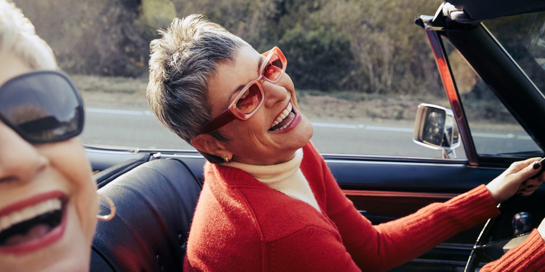 Two women sit in a convertible car with sunglasses on.