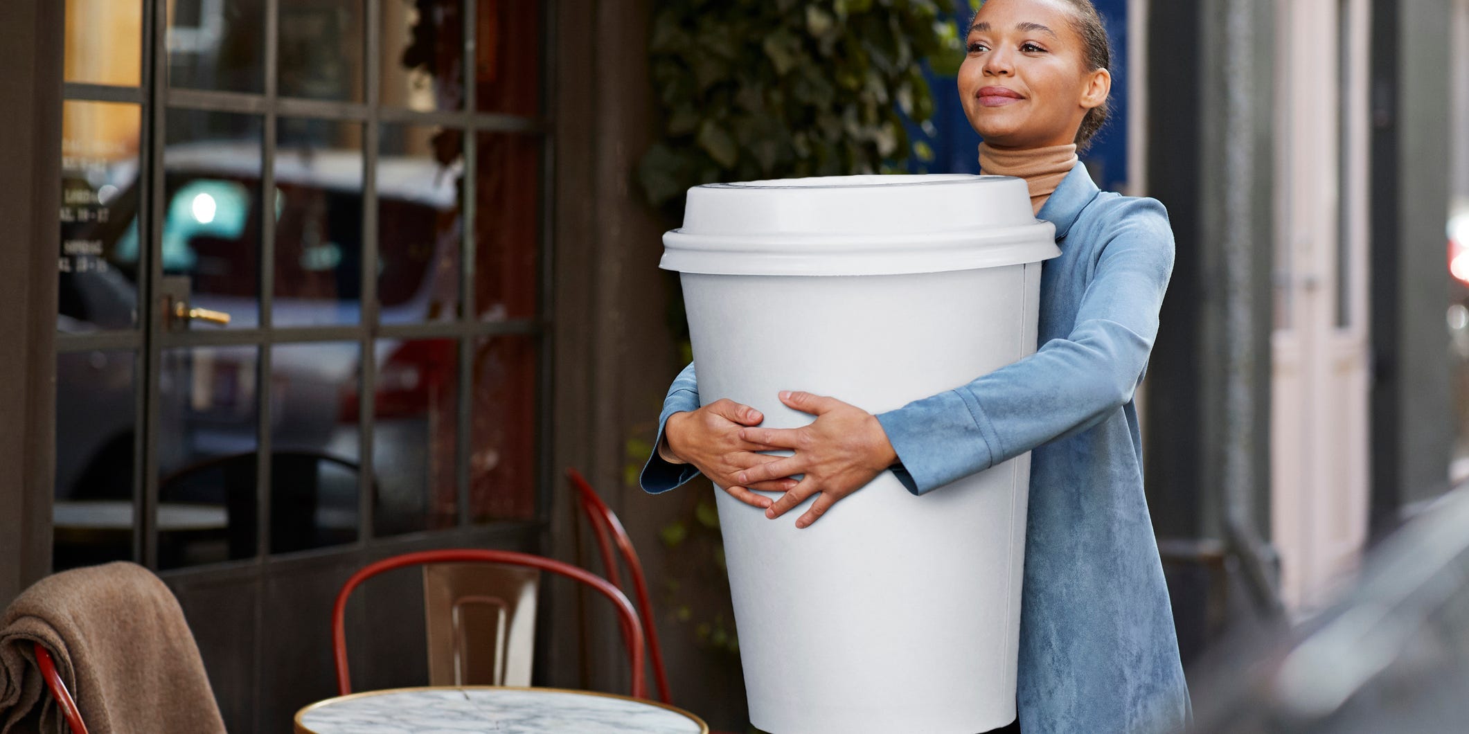 A Black woman in a denim trench coat walks down the street with a giant-sized coffee cup.