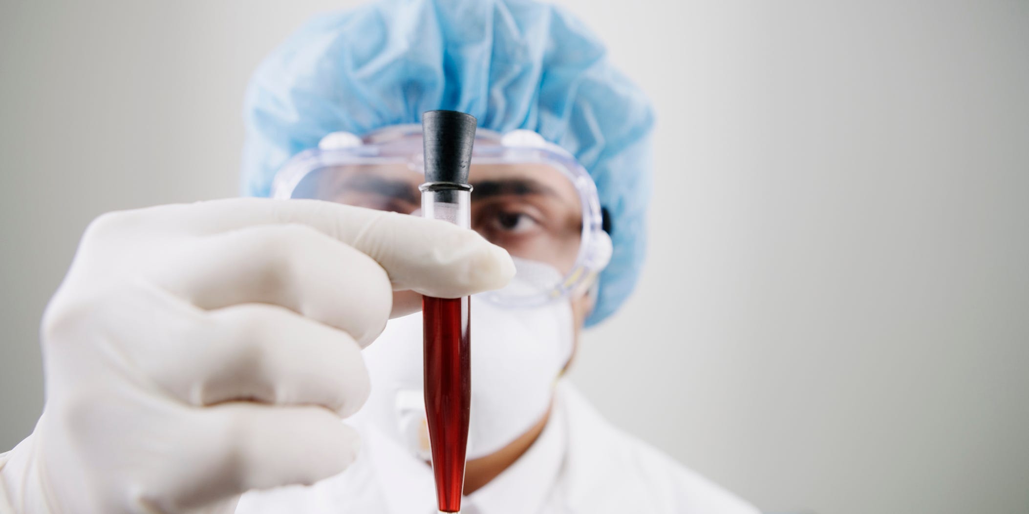 A man in a lab coat and white rubber gloves holds a vial of blood in front of the camera.