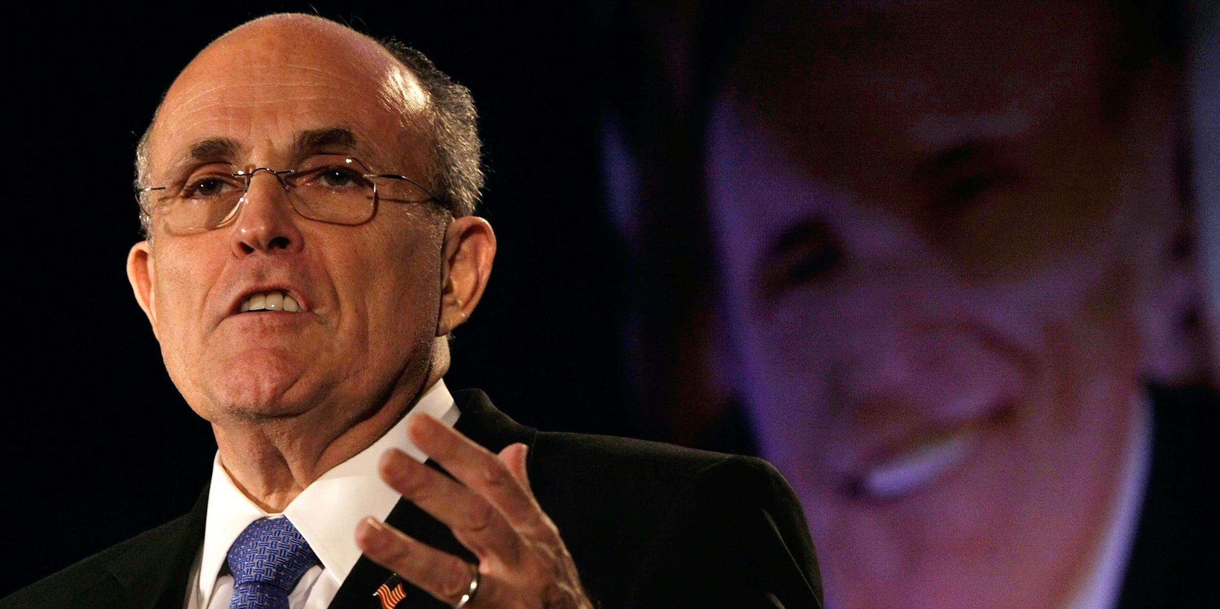 Rudy Giuliani speaks in front of a projected photo of his face.