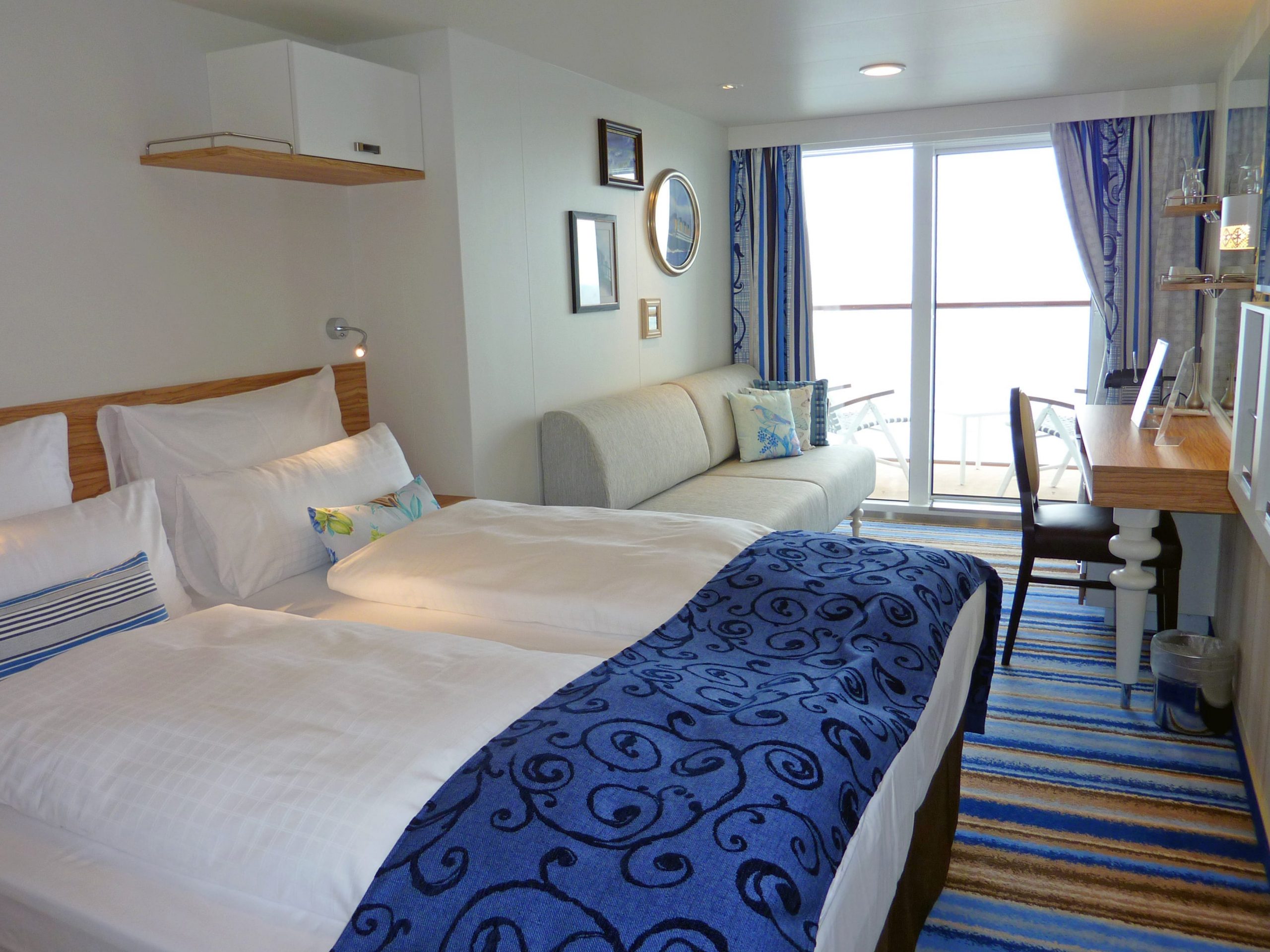 A room on a cruise ship with two beds with white sheets, a white sofa, mainly blue striped carpet, and vanity