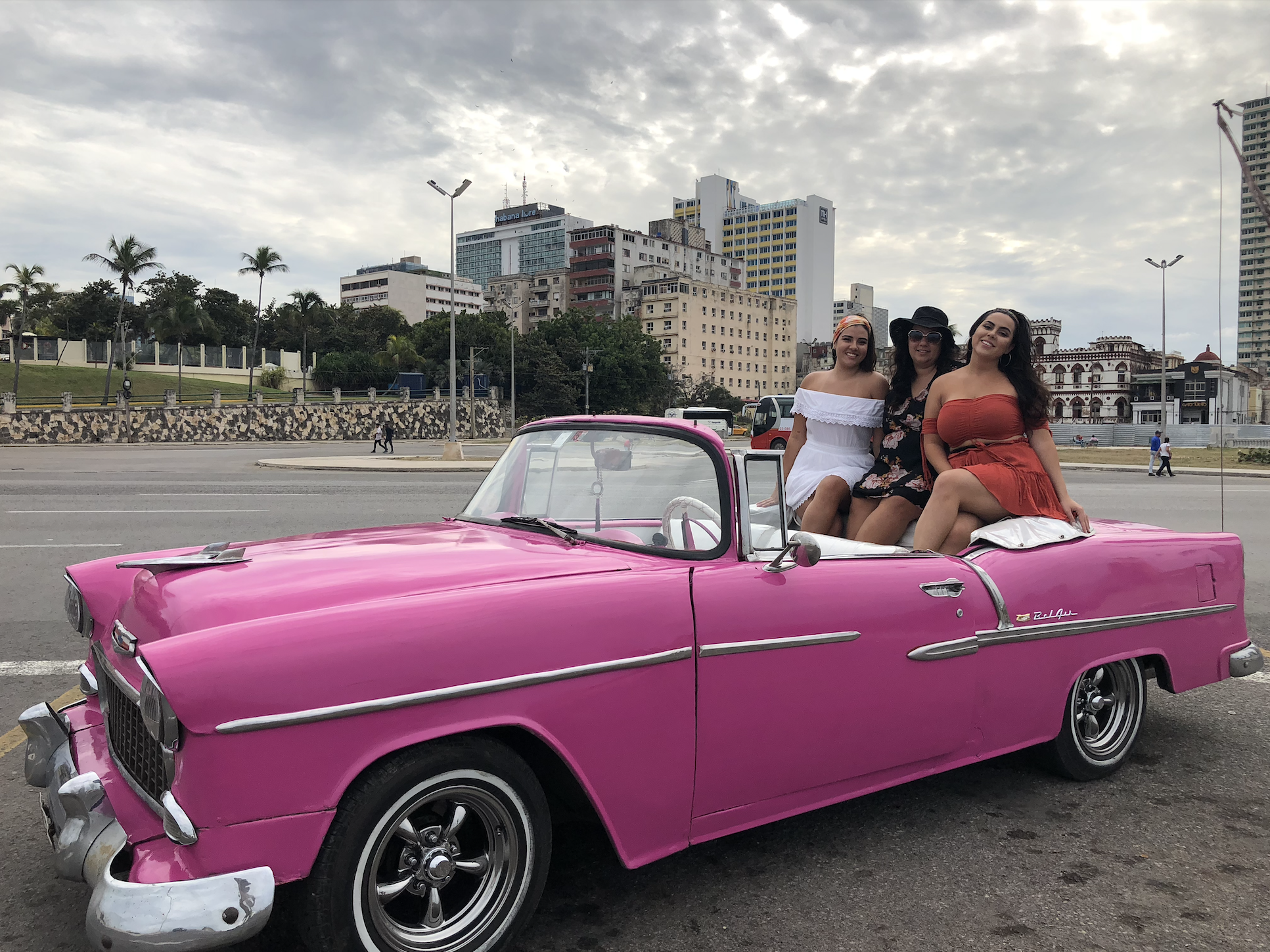 The writer and two other people sitting on the truck of a pink old-fashioned car in a city with palm trees