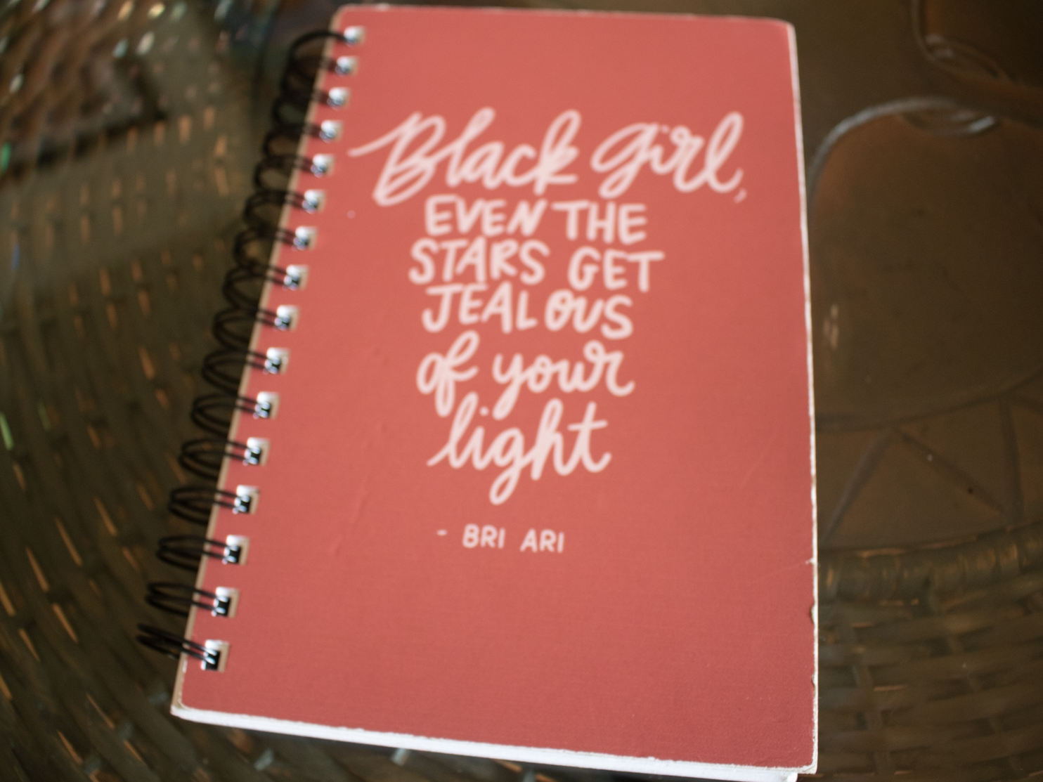 Pink travel journal on a brown table with a cover that reads "Black girl, even the stars get jealous of your light -Bri Ari"