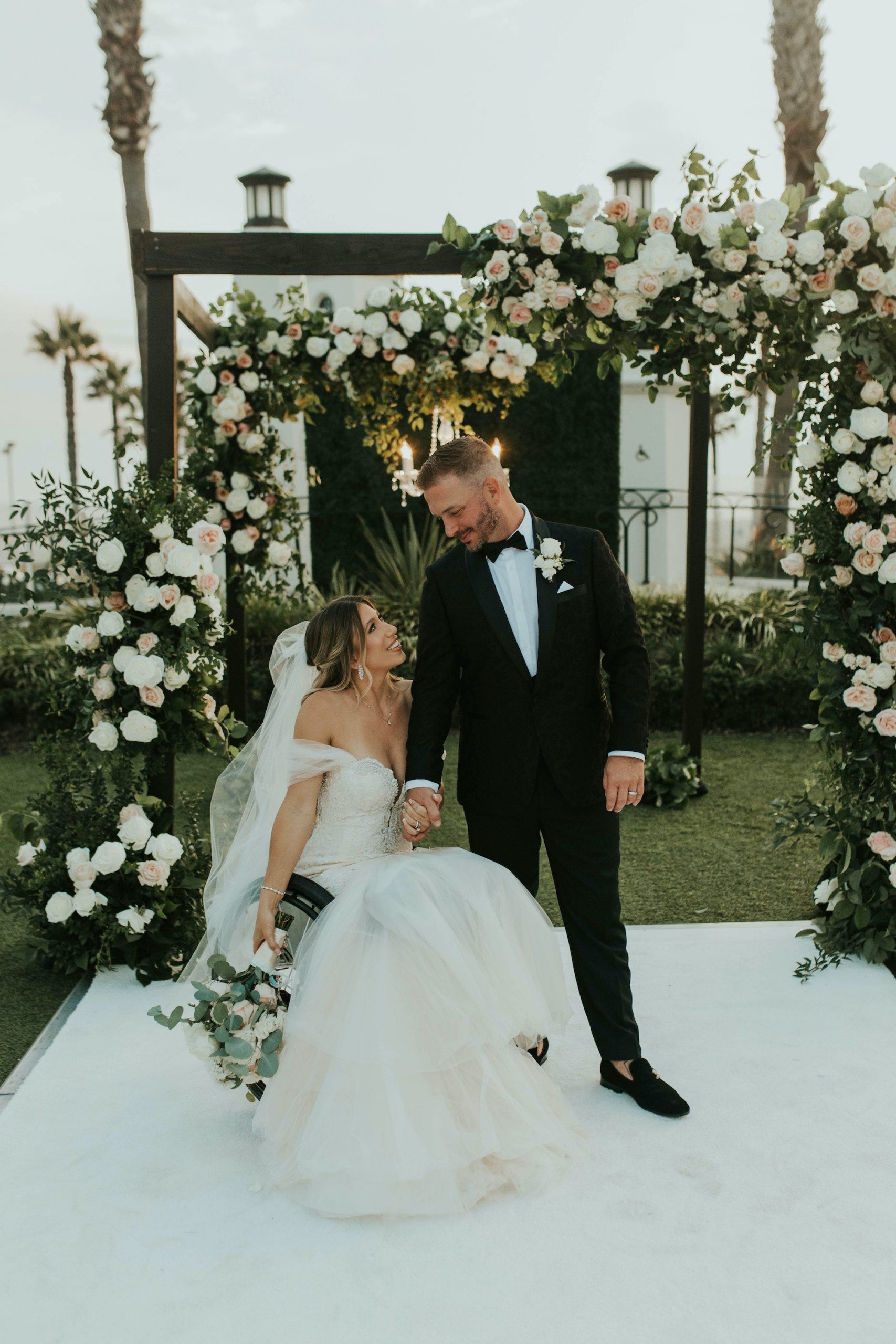 A bride sits in a wheelchair and looks up at her groom.