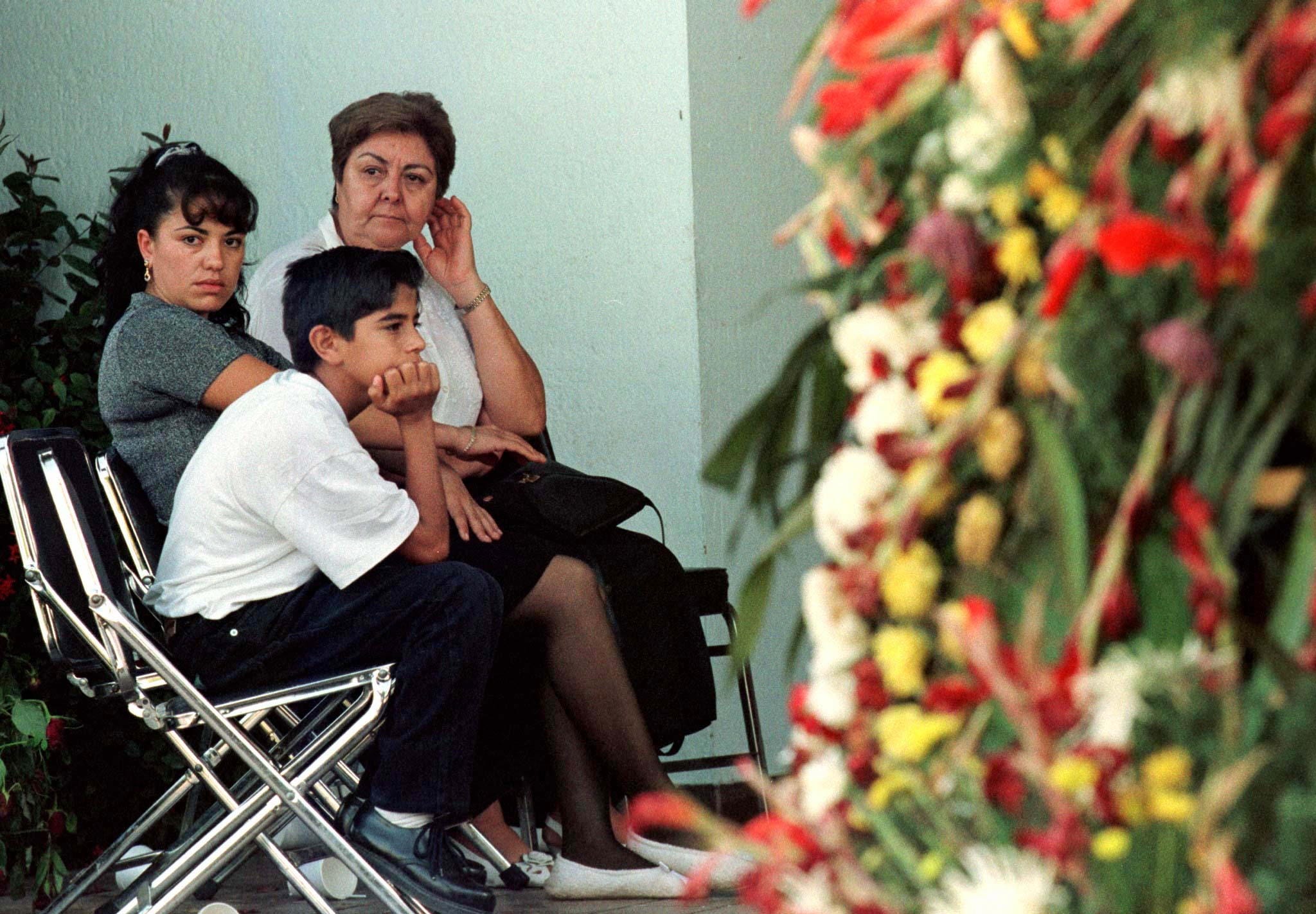 Three people sit in front of large wreaths with flowers.