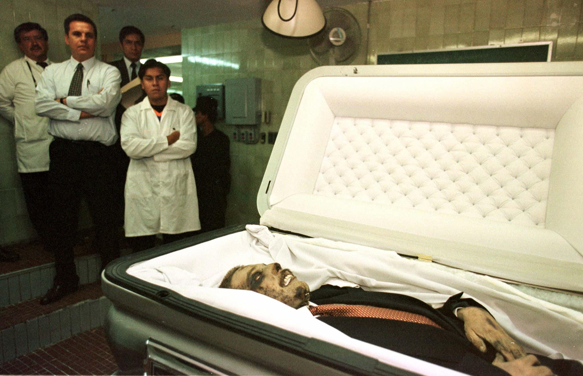 Three men hover in the background as a ghastly human body is seen in a coffin.