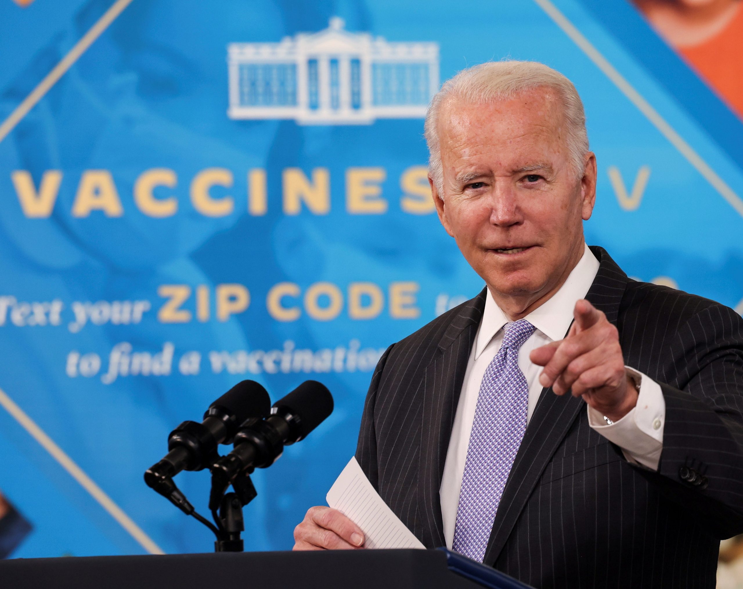 President Joe Biden stands behind microphones and points toward the camera in front of a vaccine sign