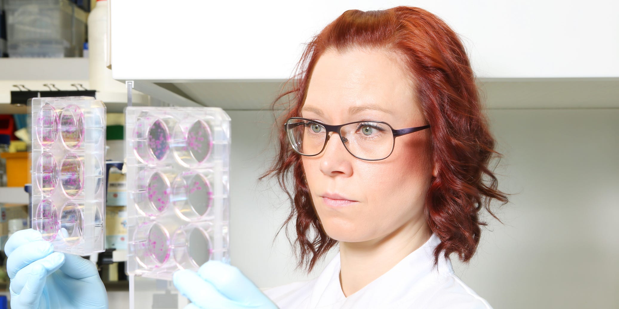 A red haired woman in a lab coat and glasses holds up two petri dishes, looking at them quizically.