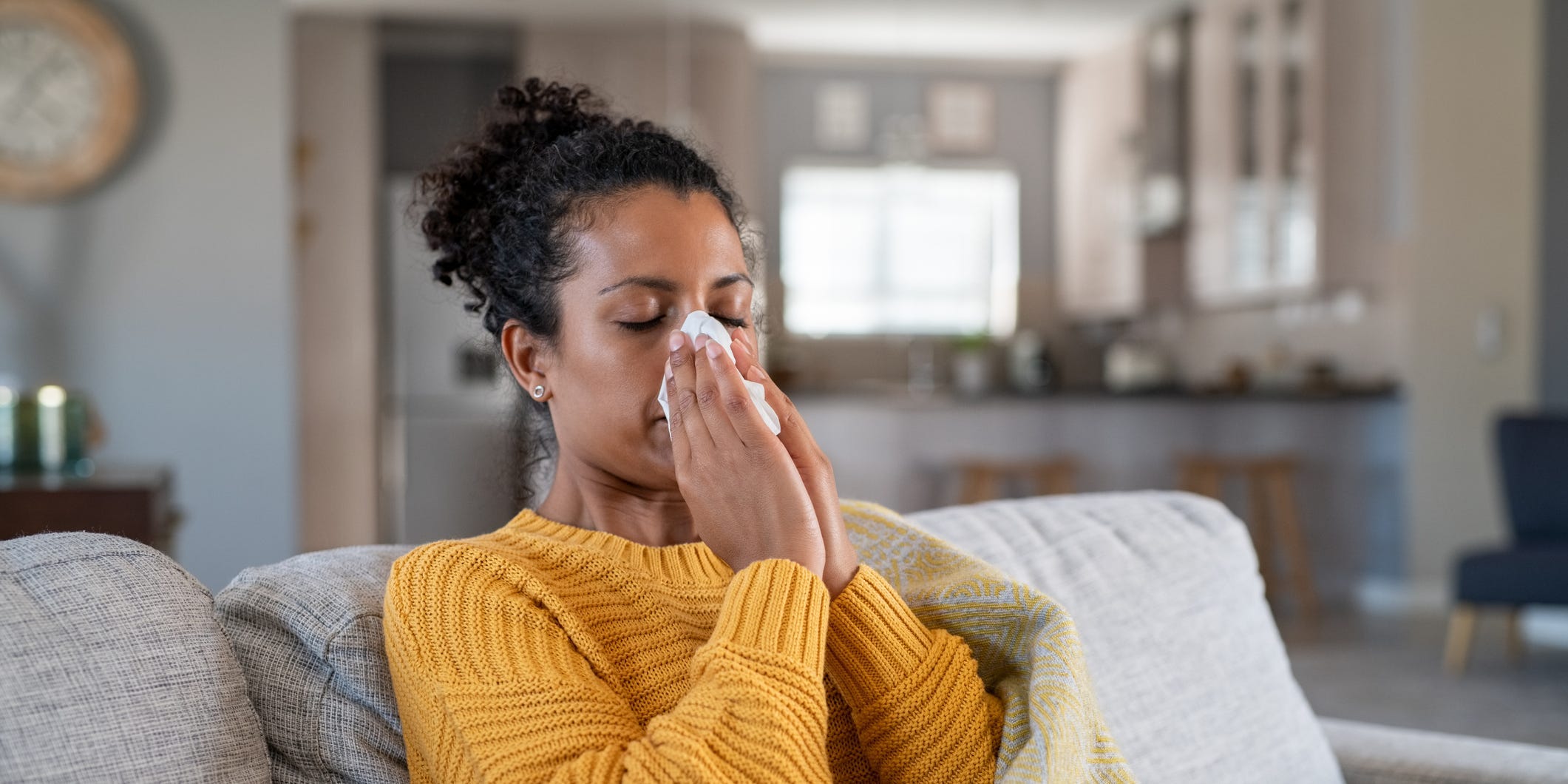 A Black woman in a yellow sweater sits on the couch wrapped in a blanket with a tissue to her nose.
