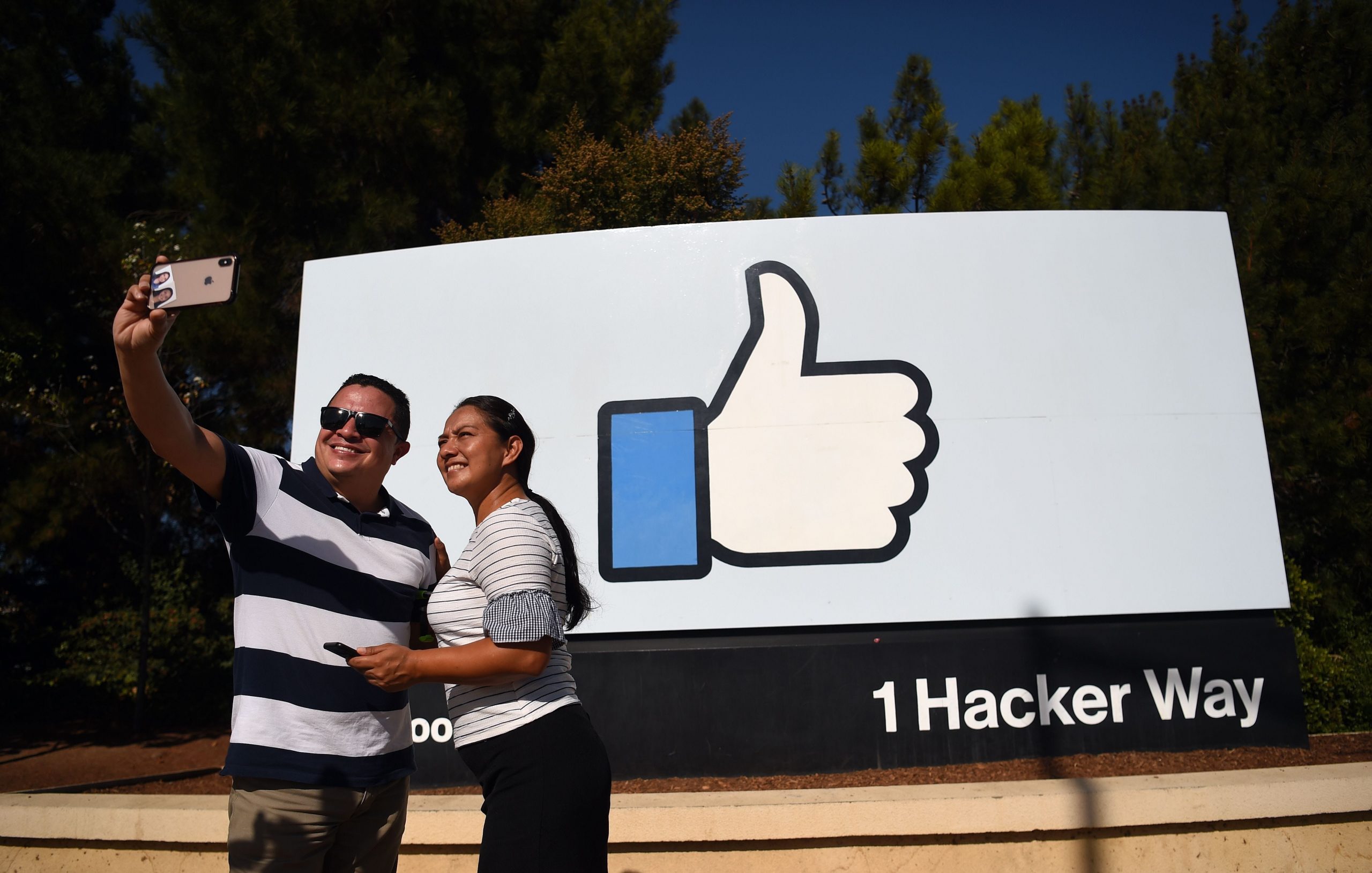 Two people taking a selfie in front of the Facebook "thumbs up" sign at the company's heaequarters