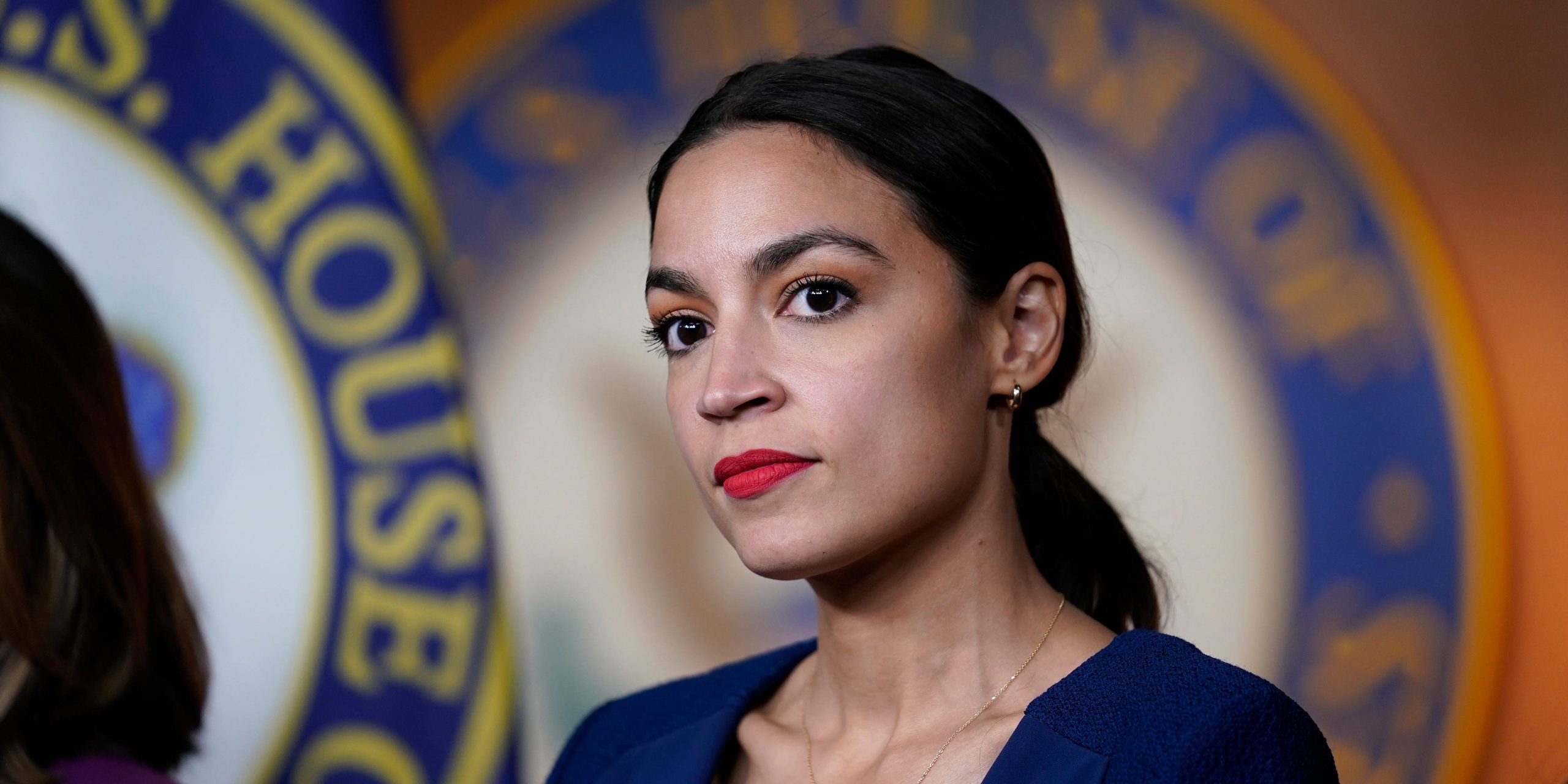 New York Congresswoman Alexandria Ocasio-Cortez in a blue blazer in front of a House of Representatives seal