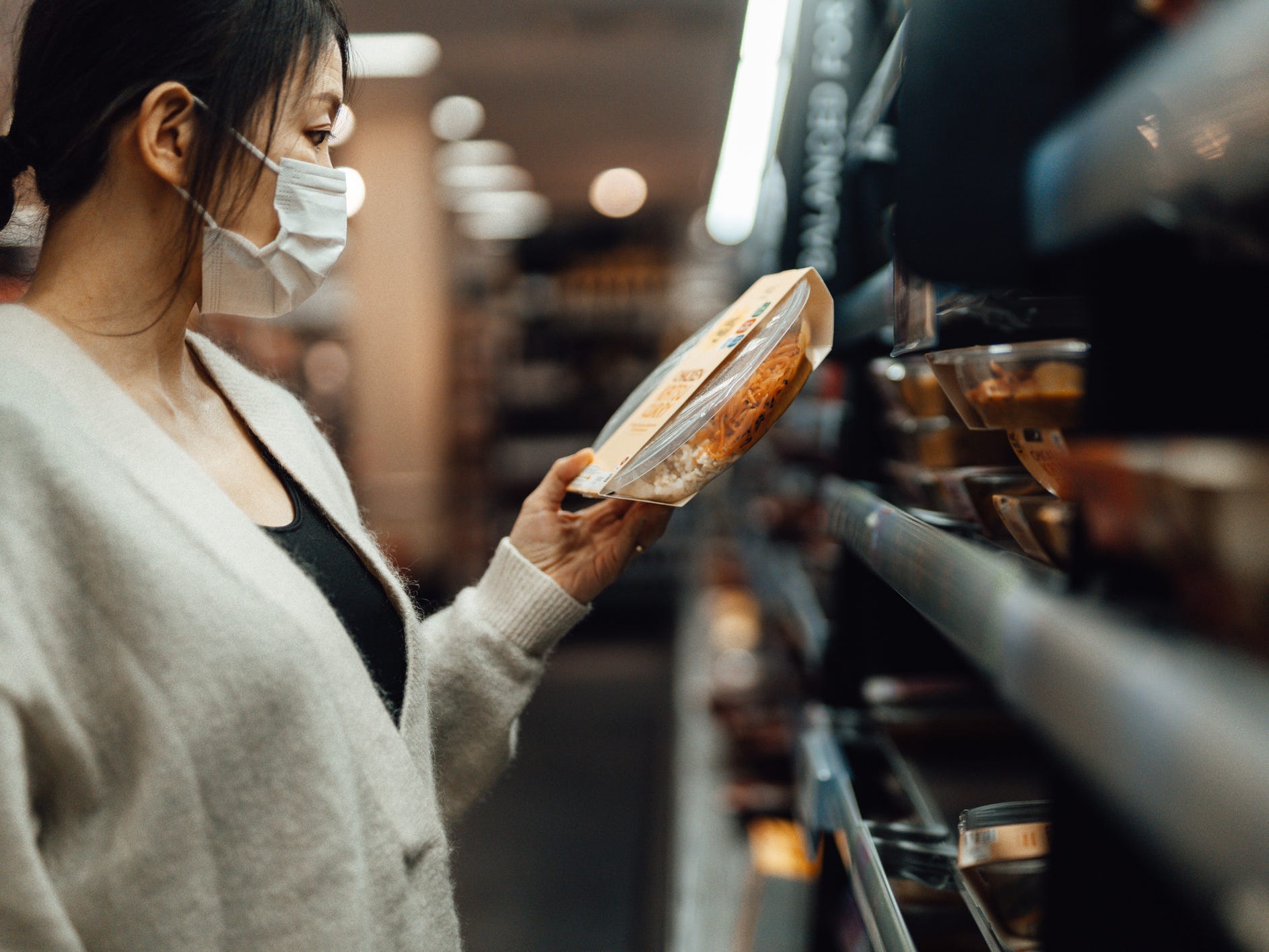 A person in a face mask holding a package of food in a grocery store aisle, looking at the nutrition label