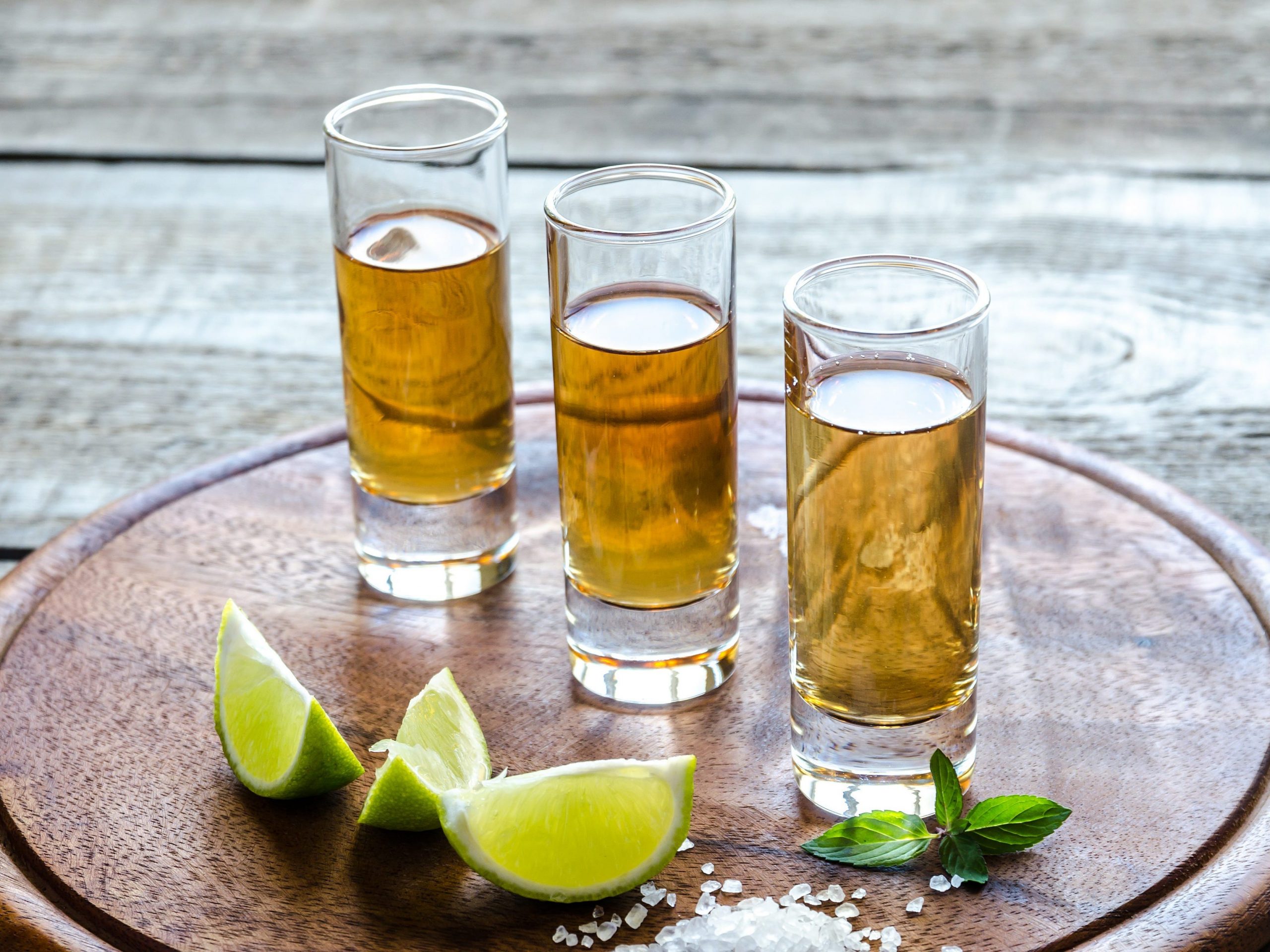 Three shots of añejo tequila on a round wooden board with lime wedges and a small pile of coarse salt