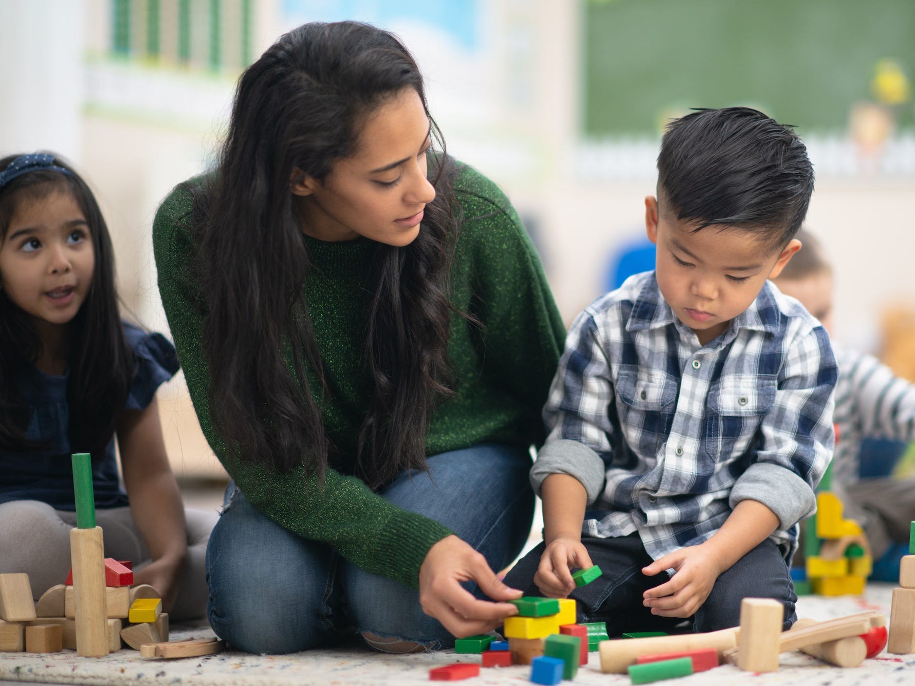 Preschool kids are playing with toy blocks and are sitting with their teacher.