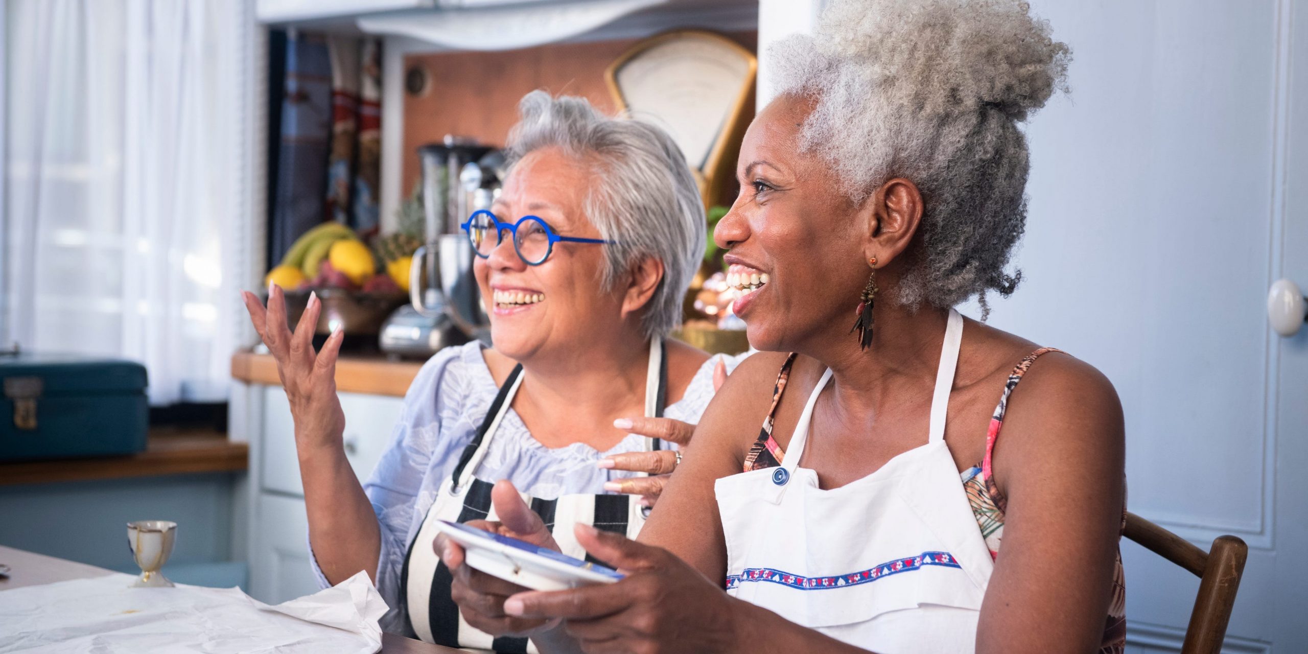 Two gray-haired women laugh together while seated at a table.