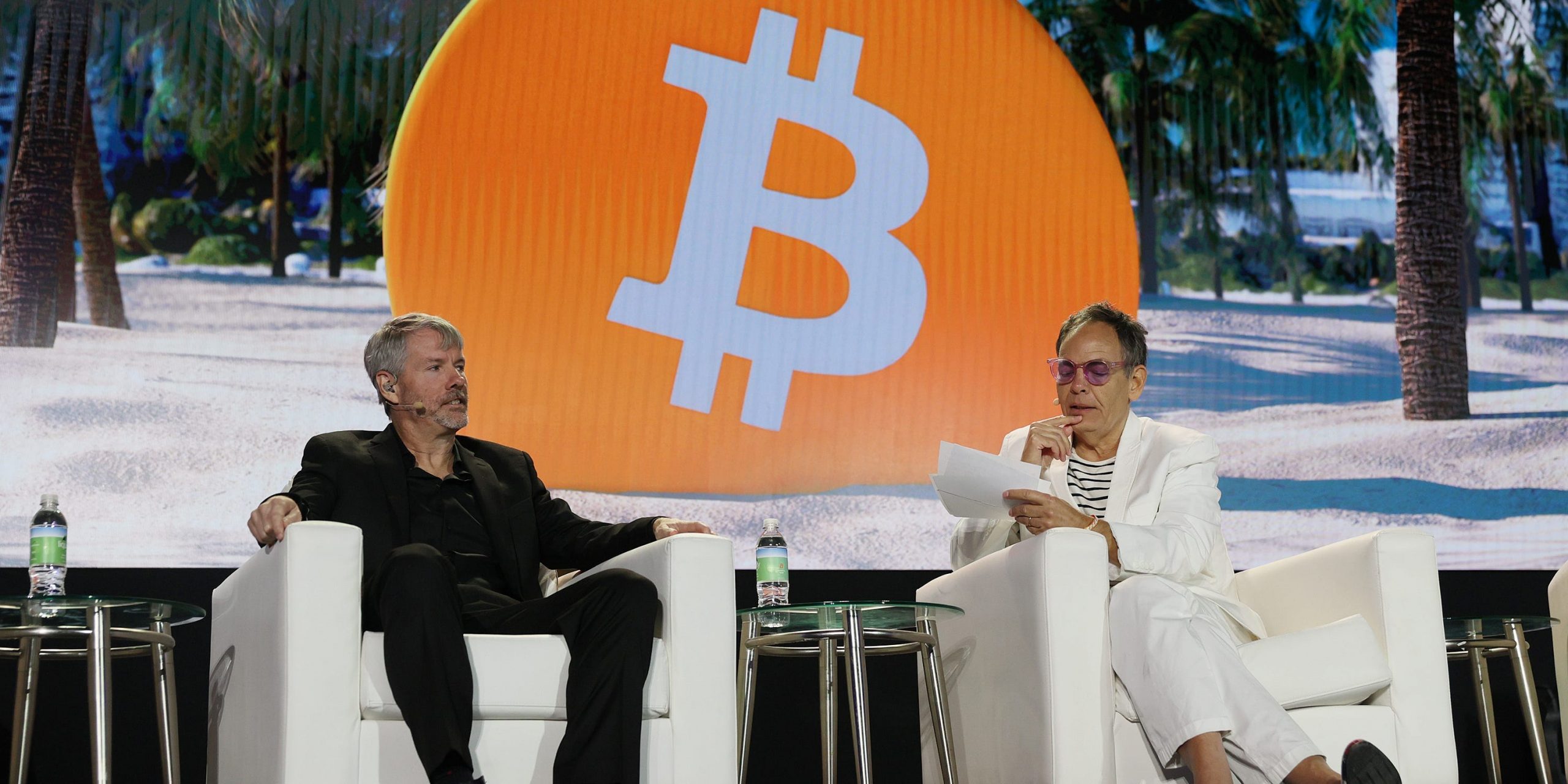 Michael saylor, dressed in all black, sits on a white couch, next to another man in all white, with a giant orange Bitcoin logo behind them.