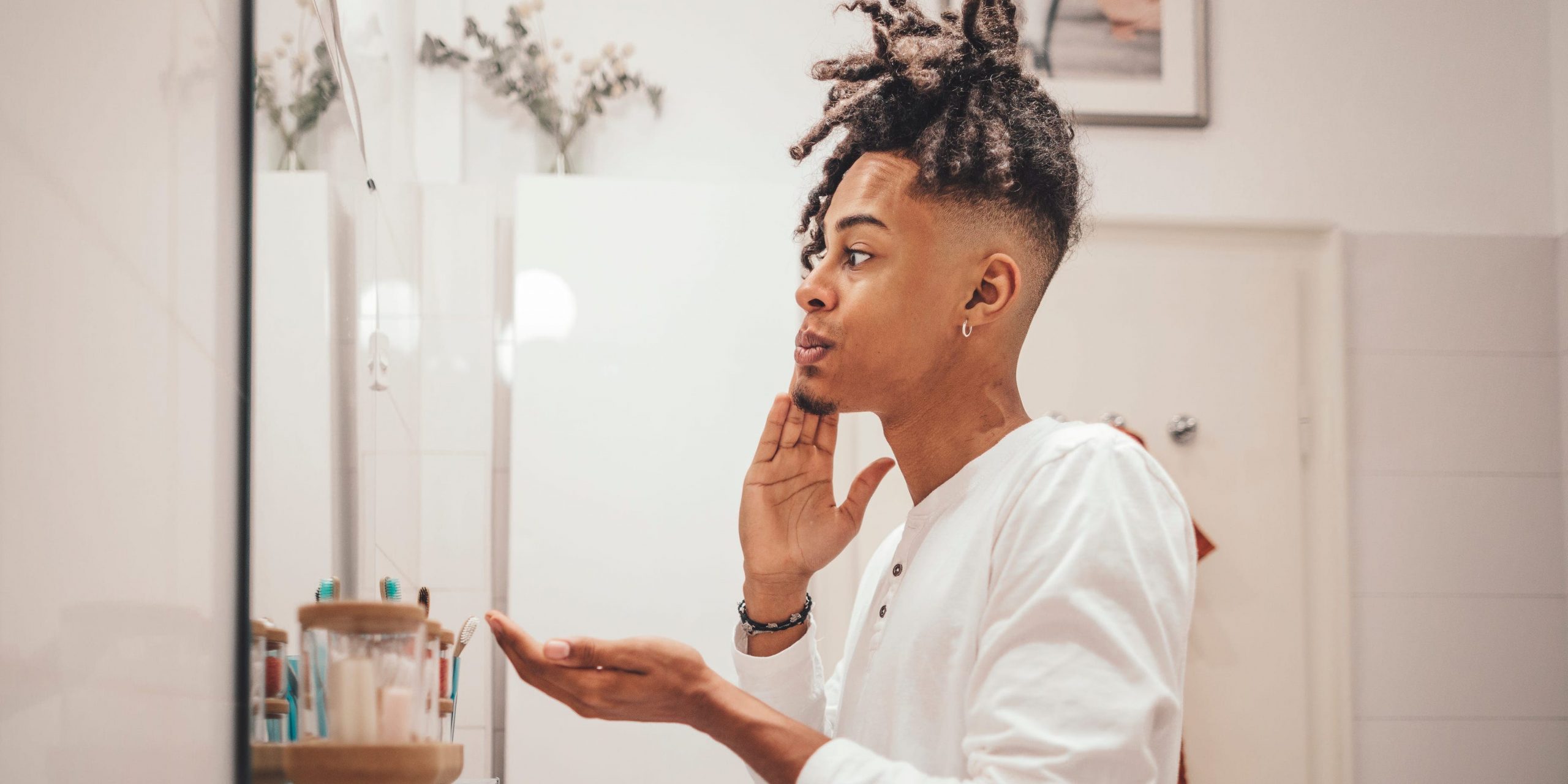 A young man in profile looks in the mirror as he applies skincare product to his face.