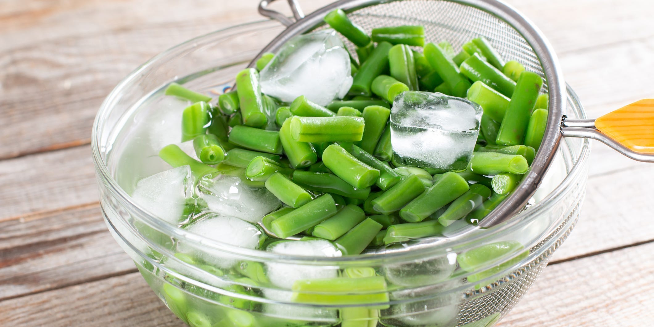 A strainer pulling cut green beans out of a bowl of ice water