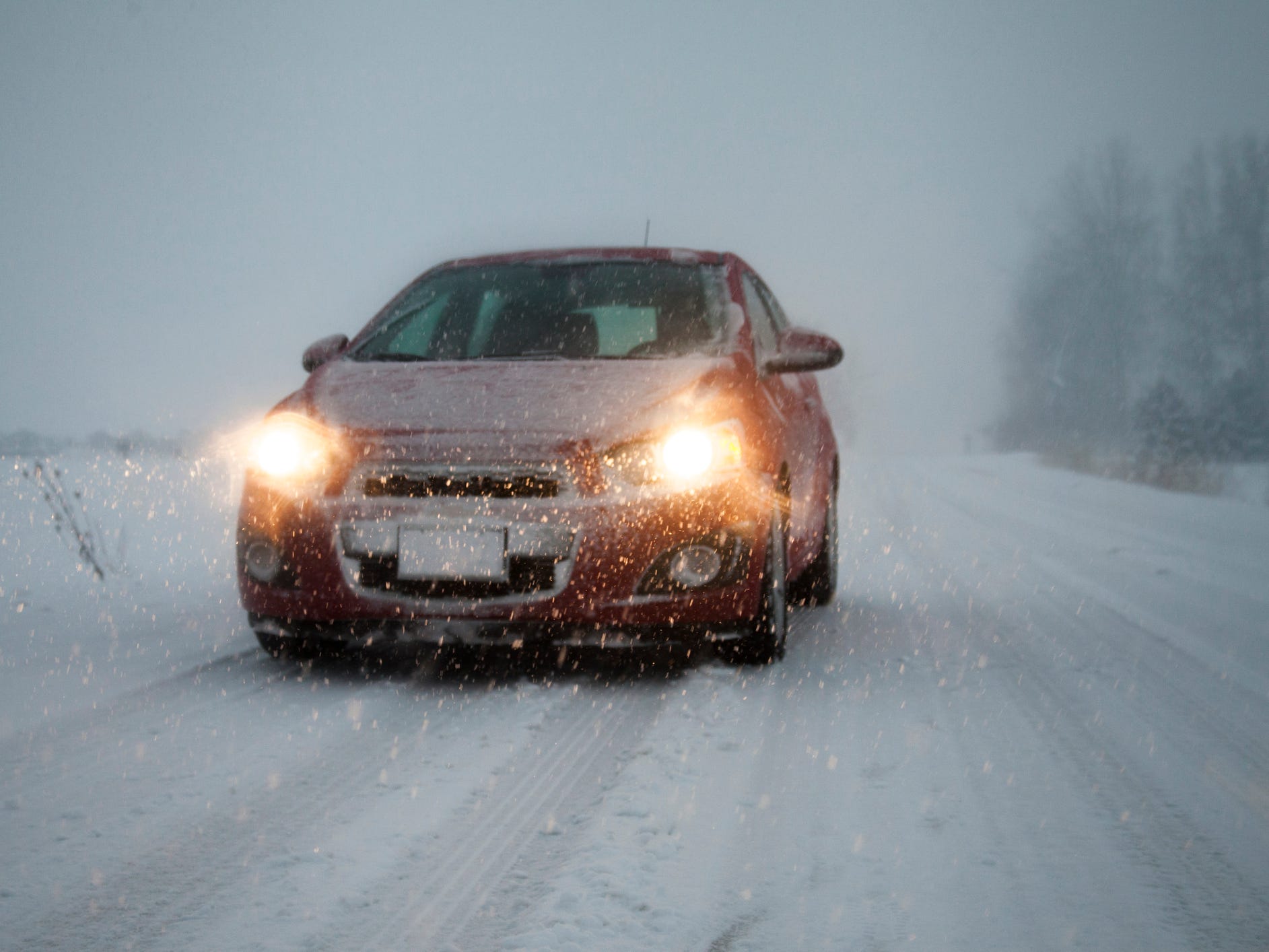 A red car with its headlights on driving in the snow