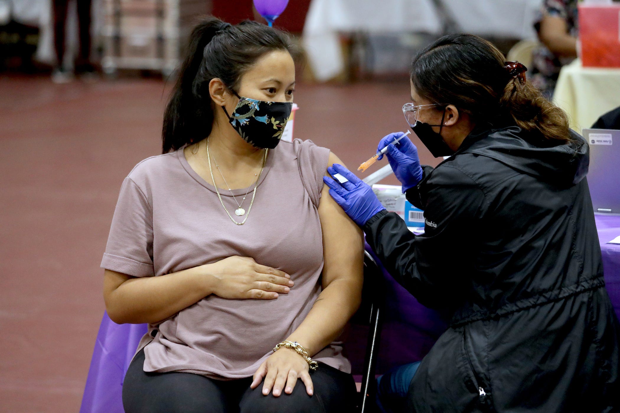 a pregnant woman, sitting and clutching her belly, getting a covid 19 booster shot stuck into her arm.