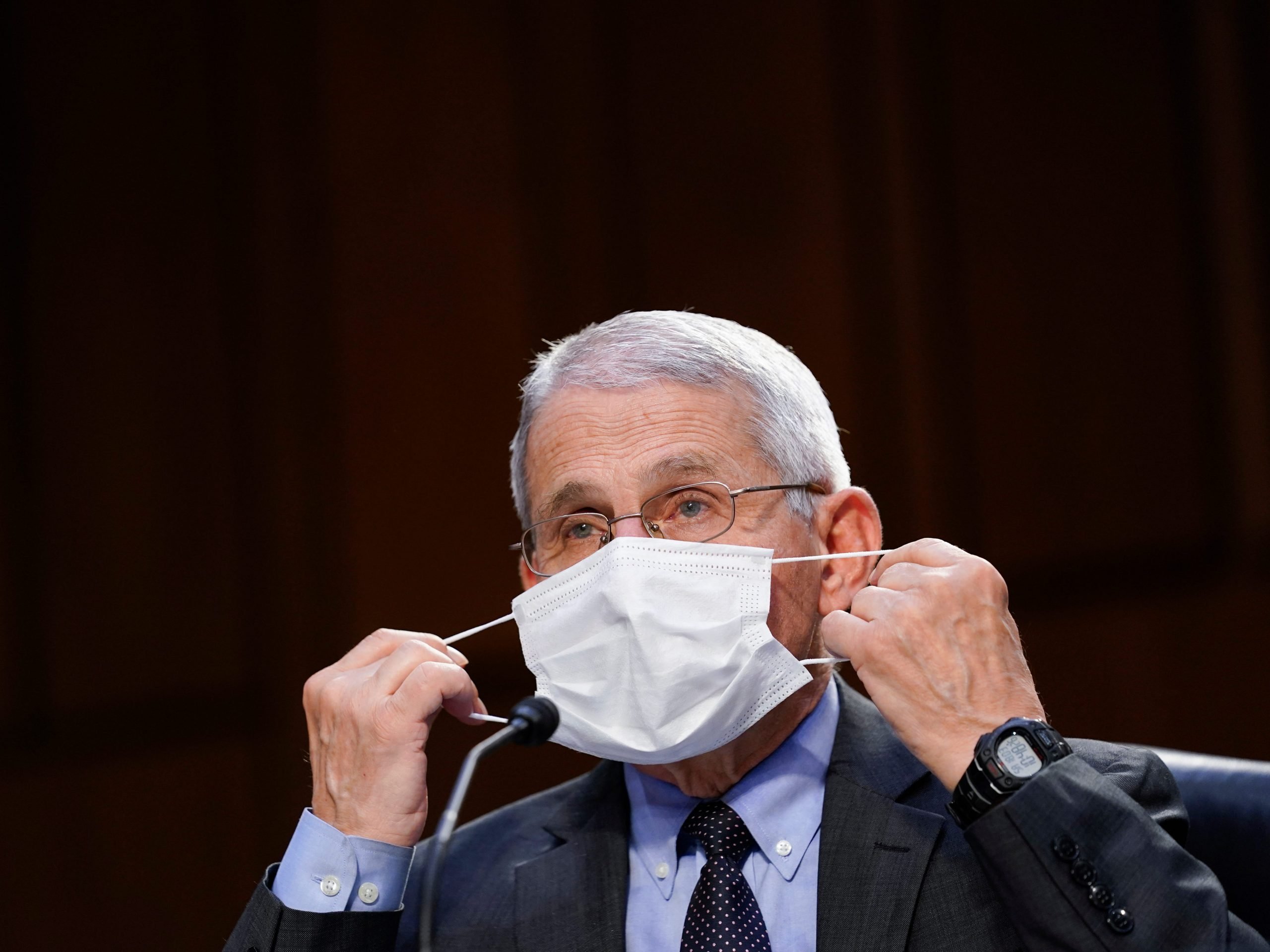 Dr. Anthony Fauci puts on white mask, dressed in suit. Black background.