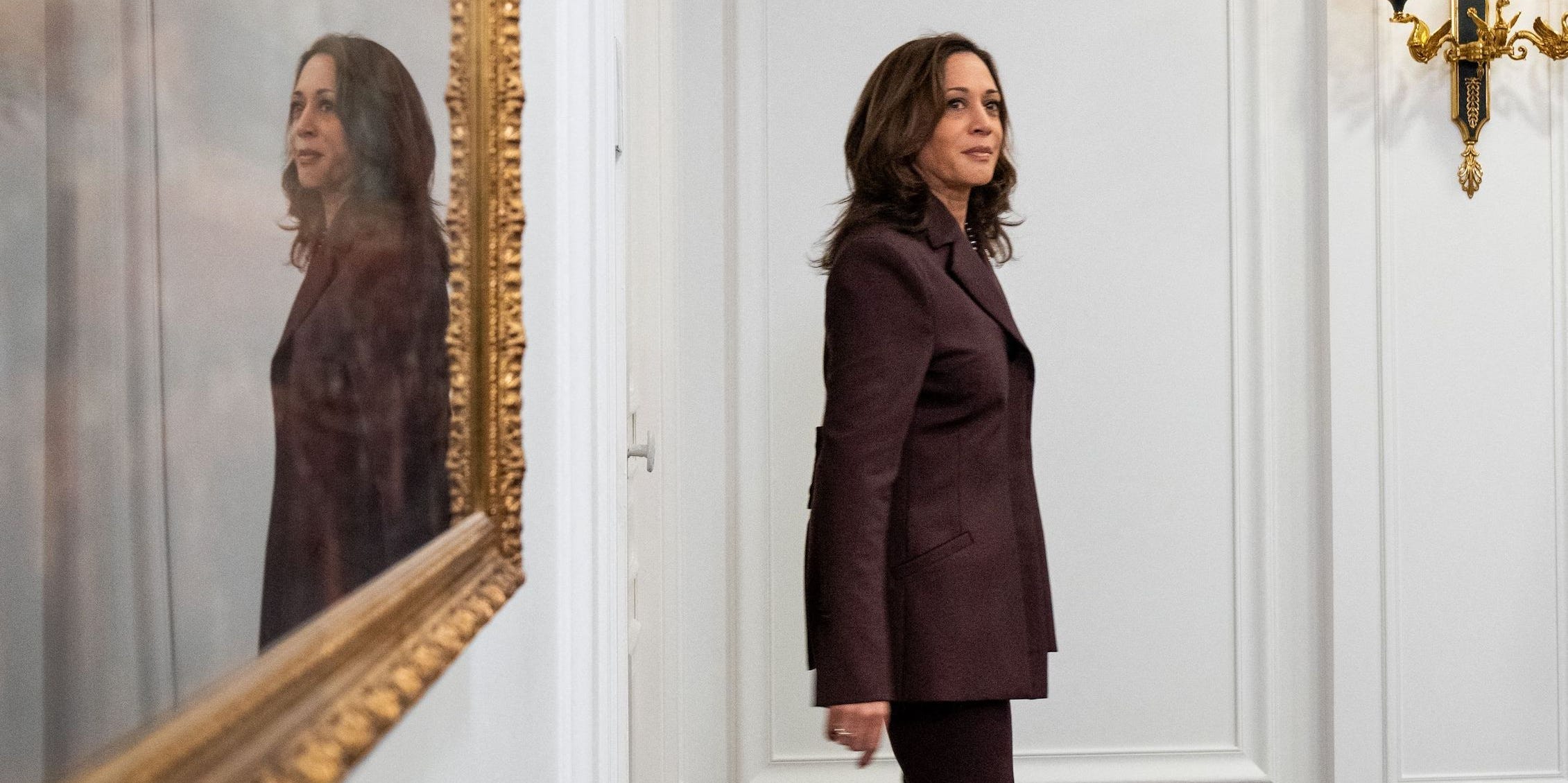 Vice President Kamala Harris walks into a room with her reflection on a mirror behind her.