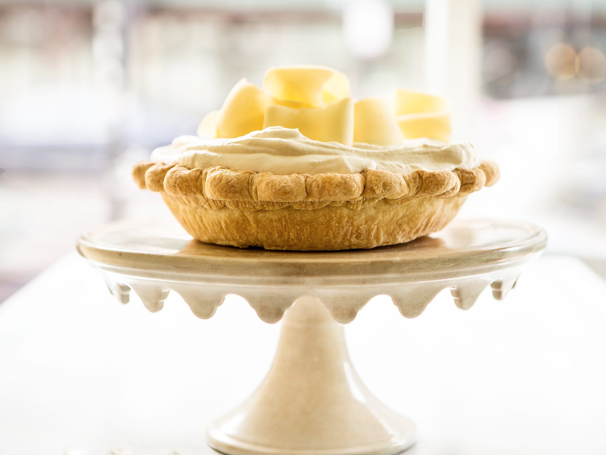 A cream pie topped with white chocolate ribbons on a white cake stand