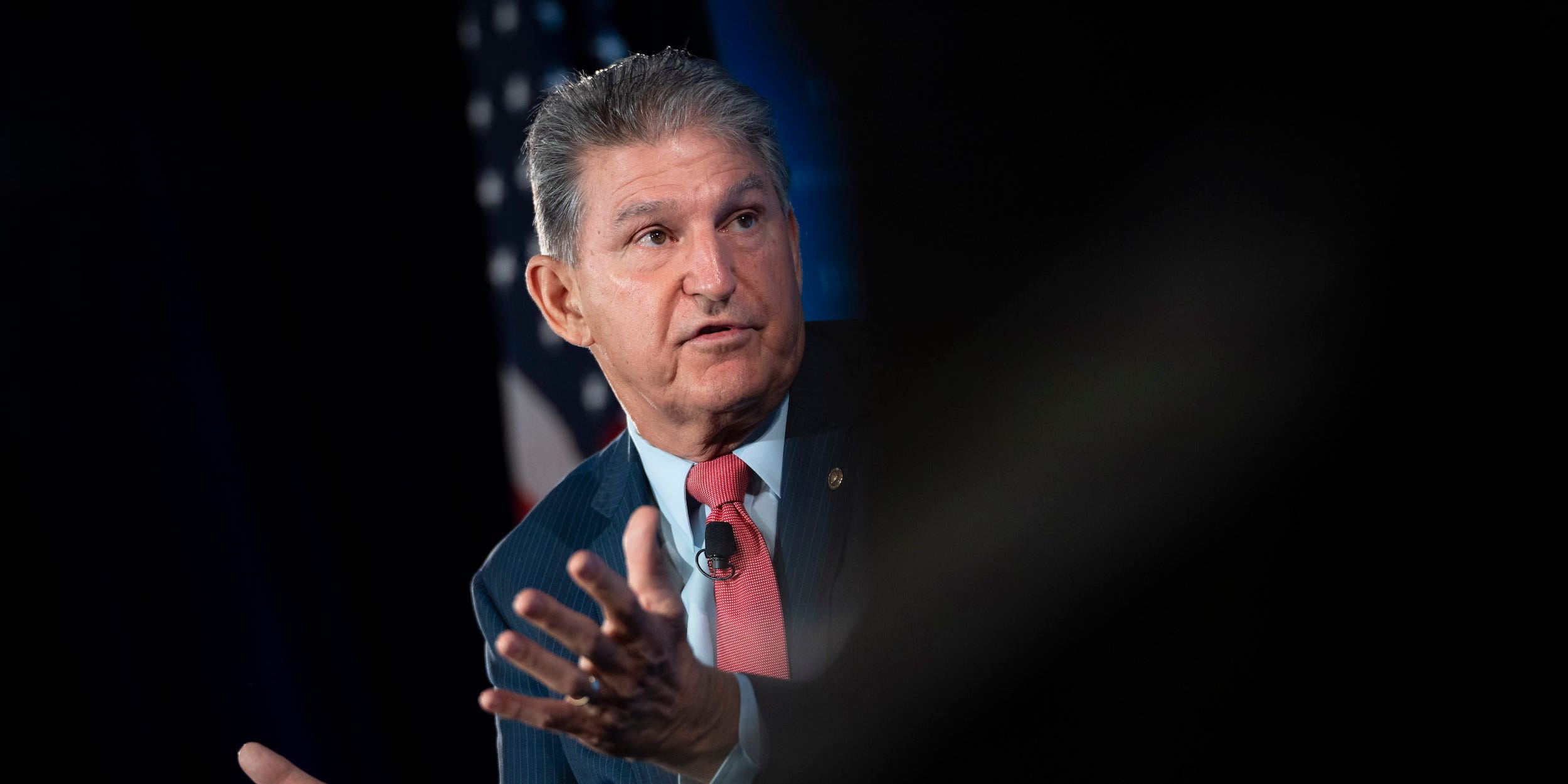 Senator Joe Manchin gestures in front of an American flag with his palms up, the right half of the photo is blurred out in black.