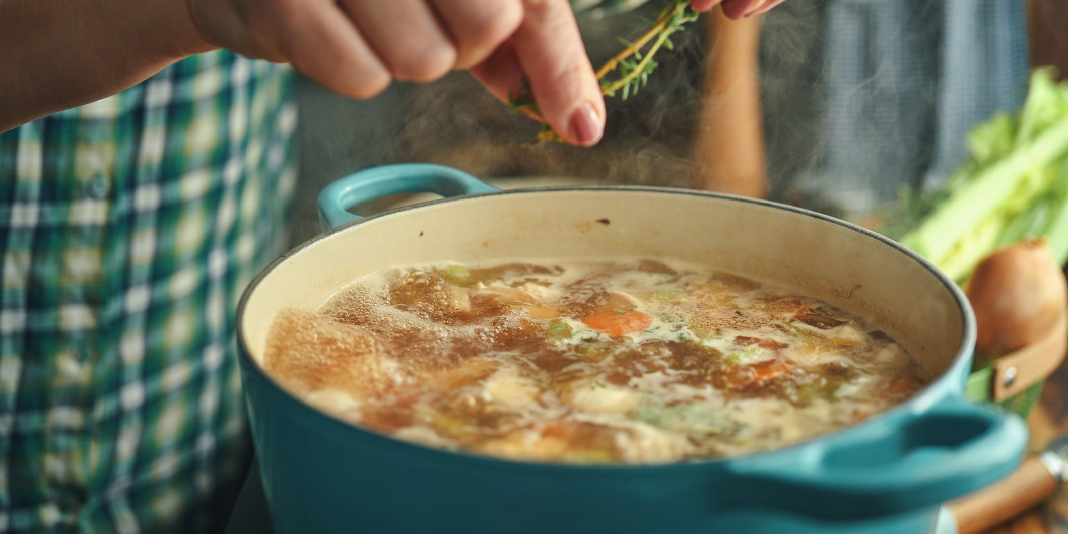 A person's hands adding fresh thyme to a boiling pot of chicken soup