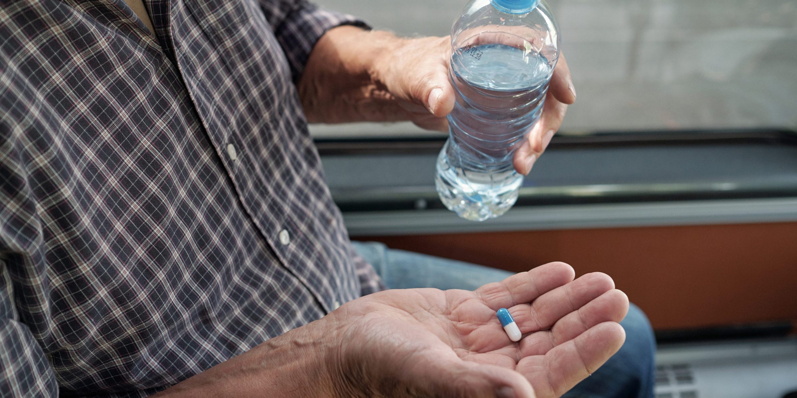 Close-up of unrecognizable aged man in casual shirt holding bottle of water and taking dramamine pill in bus