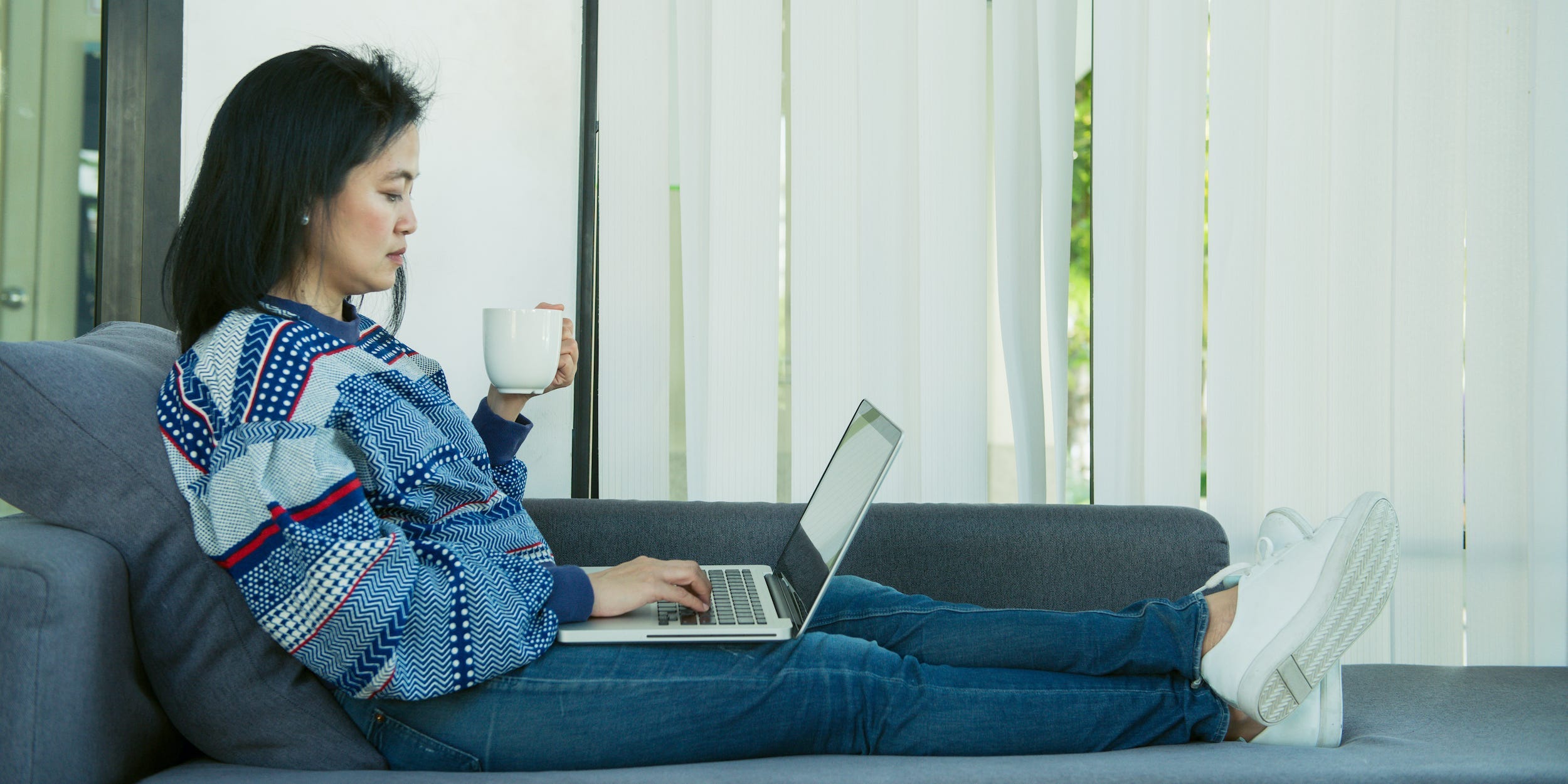 woman using laptop computer on couch at home with coffee in hand