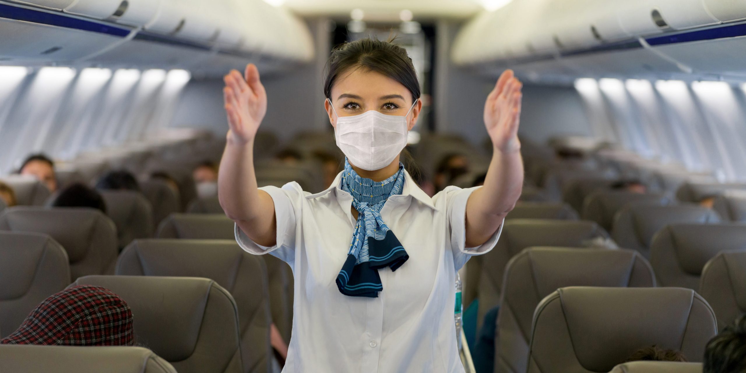 A woman flight attendants wearing a mask gestures towards the exits on an airplane.