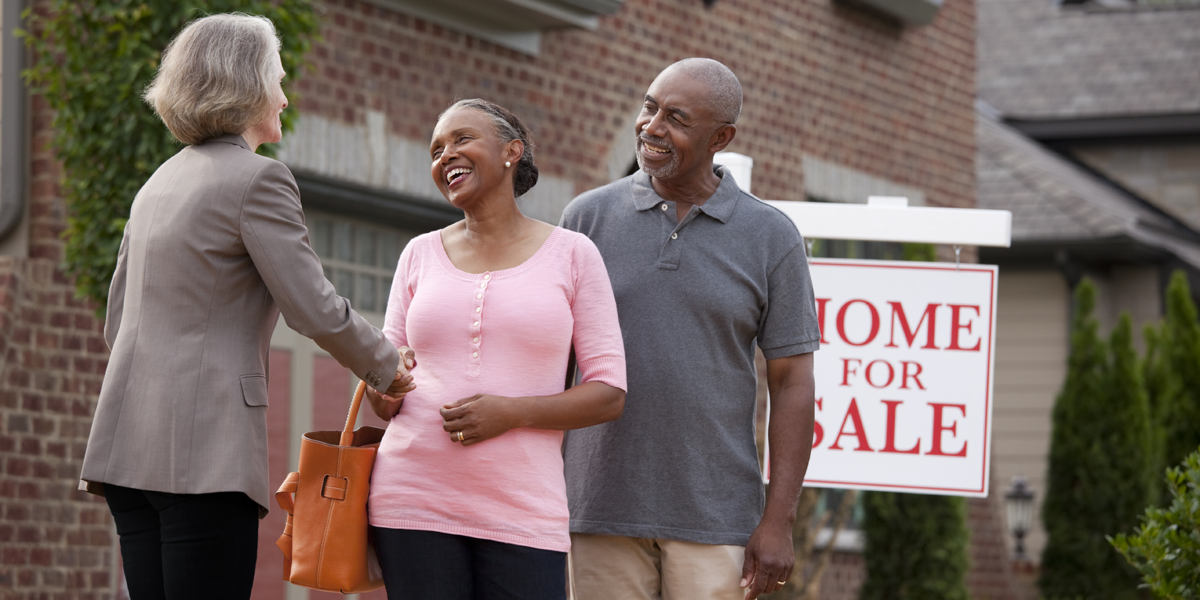 Realtor shaking hands with smiling couple outside house for sale.