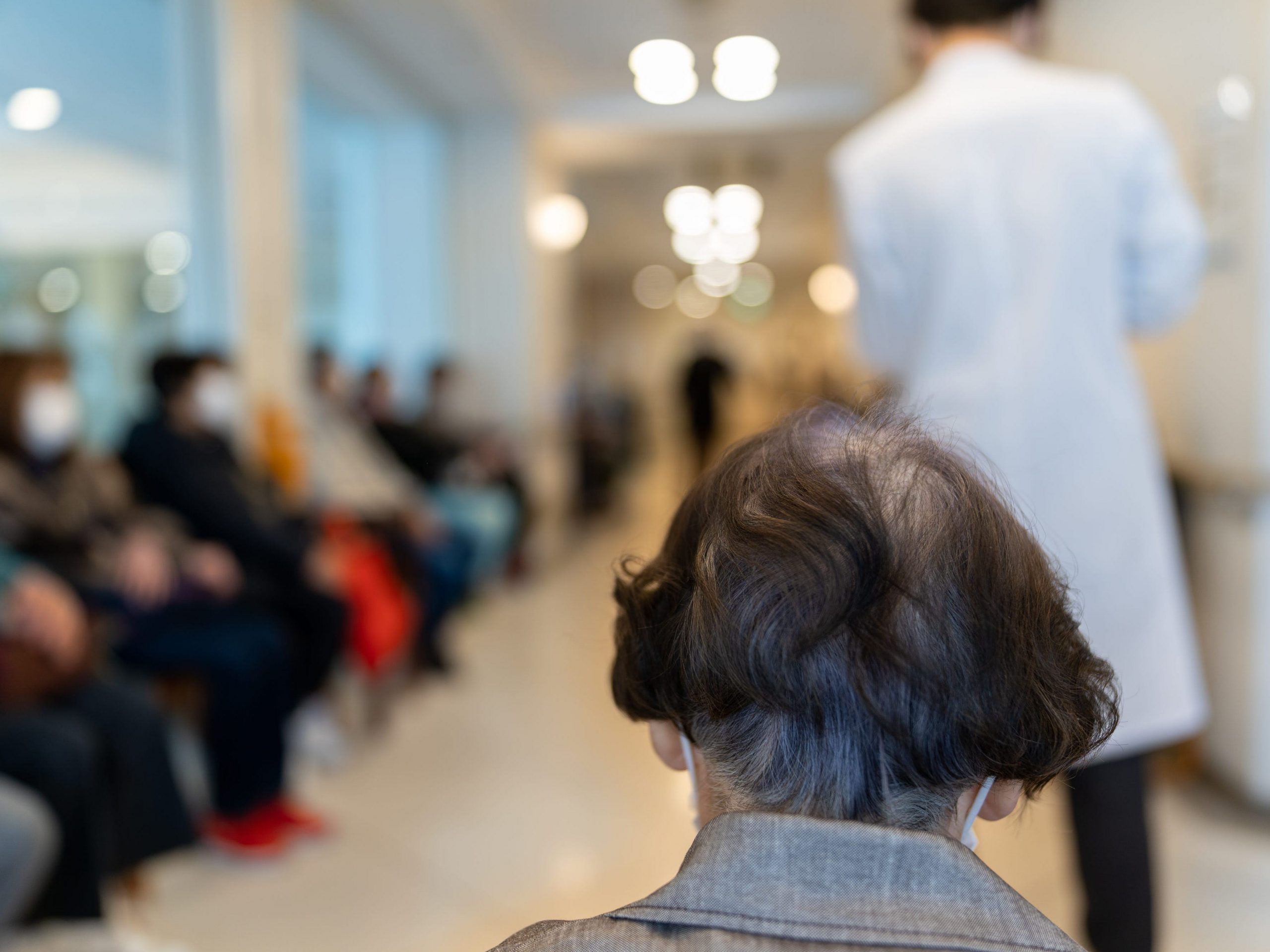 Japanese senior woman wearing face mask sitting in a hospital lobby as patients wait to her side and a doctor stands in front.