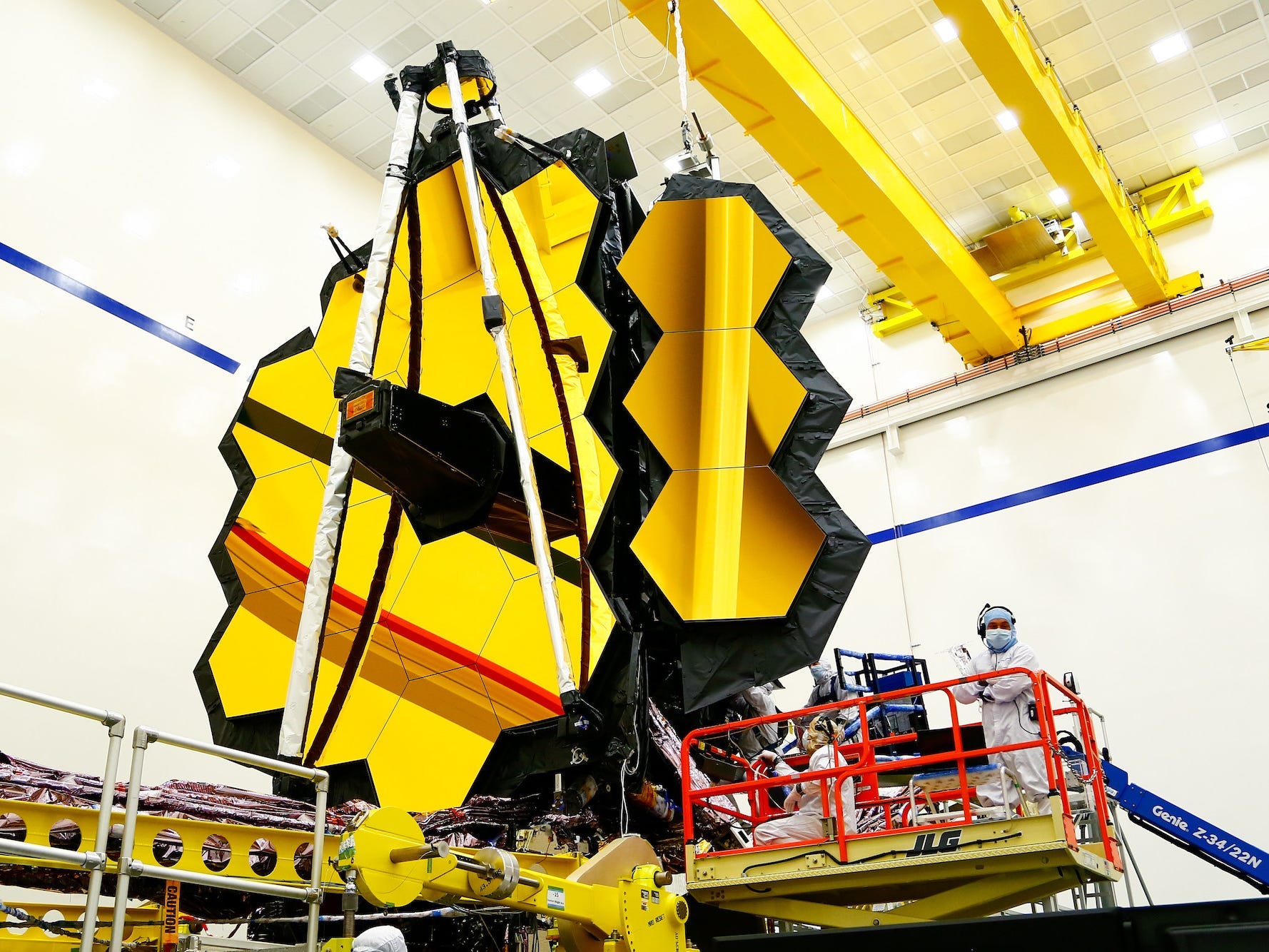 The panels of the James Webb space telescope are pictured supported by a harness behind people wearing protective suits.