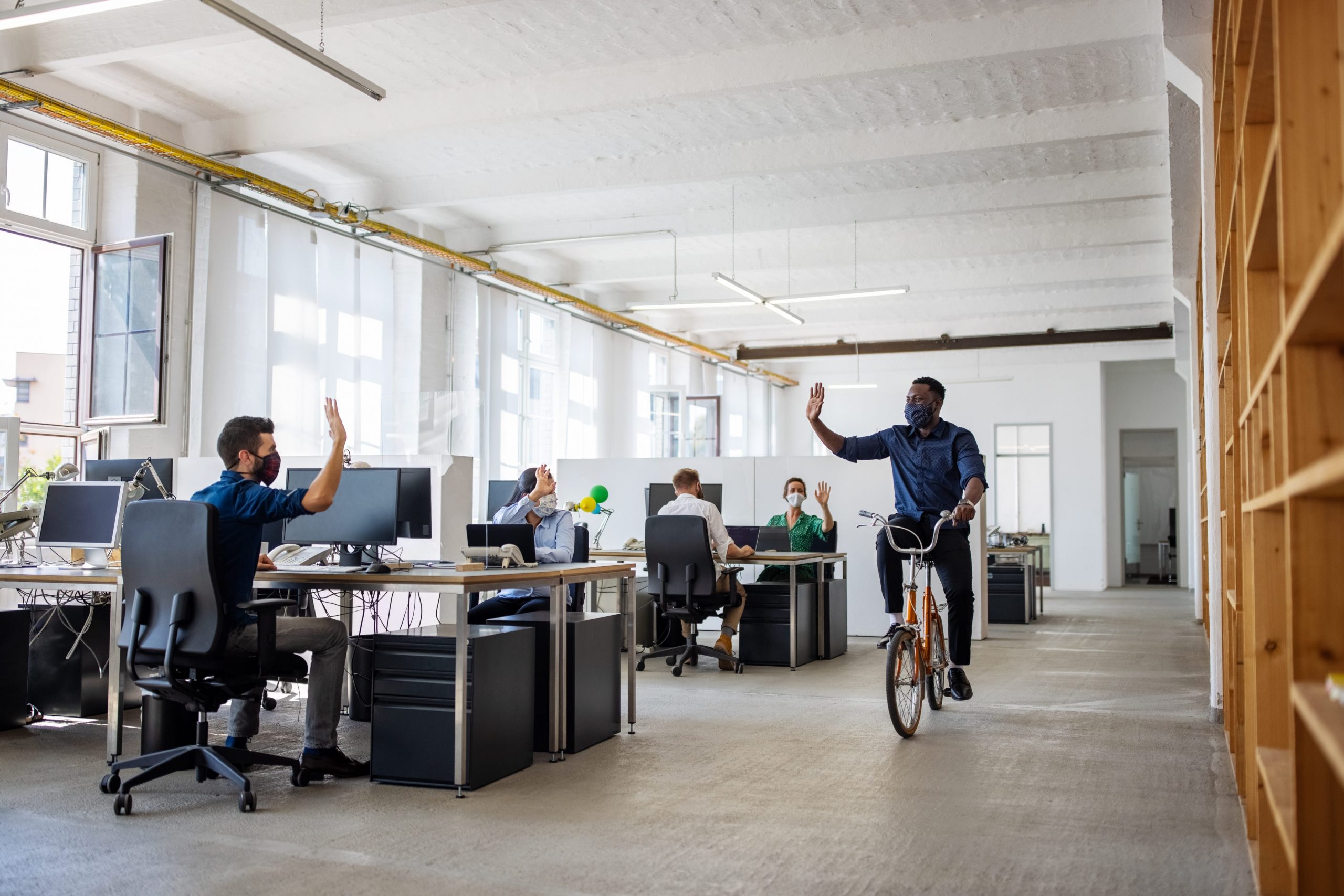 A man ride a bicycle through an office while waving to his coworker.