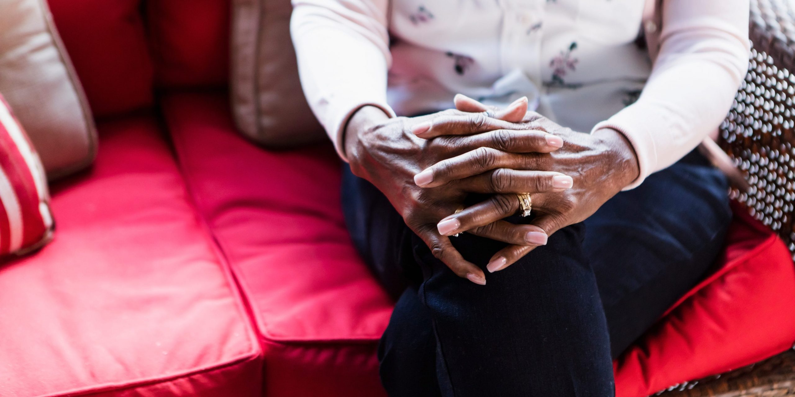 A older woman sits on a couch and clasps her hands over her knees with her legs crossed.