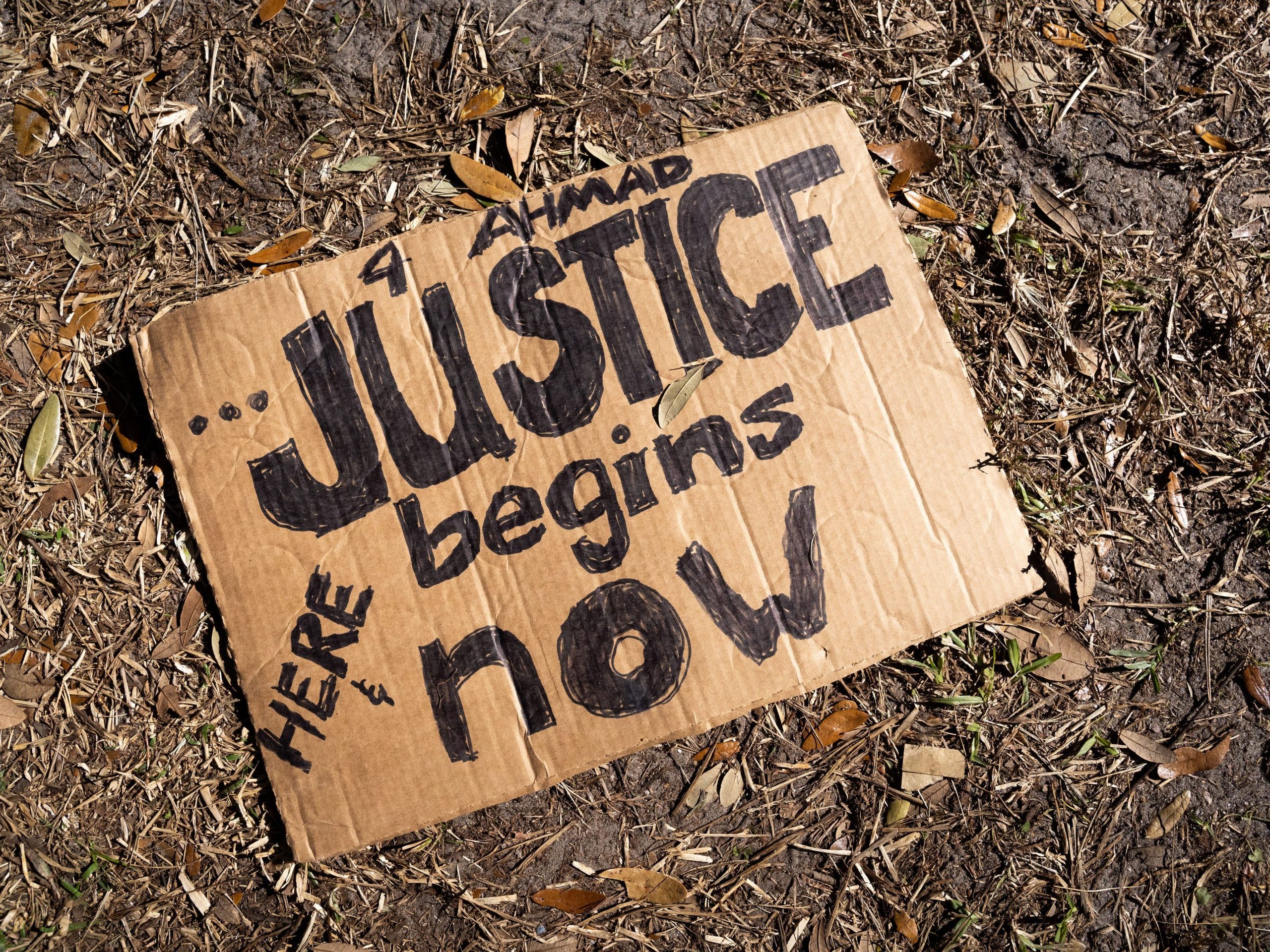 Cardboard sign on the dirt ground that reads '4 Ahmad, Justice begins now."