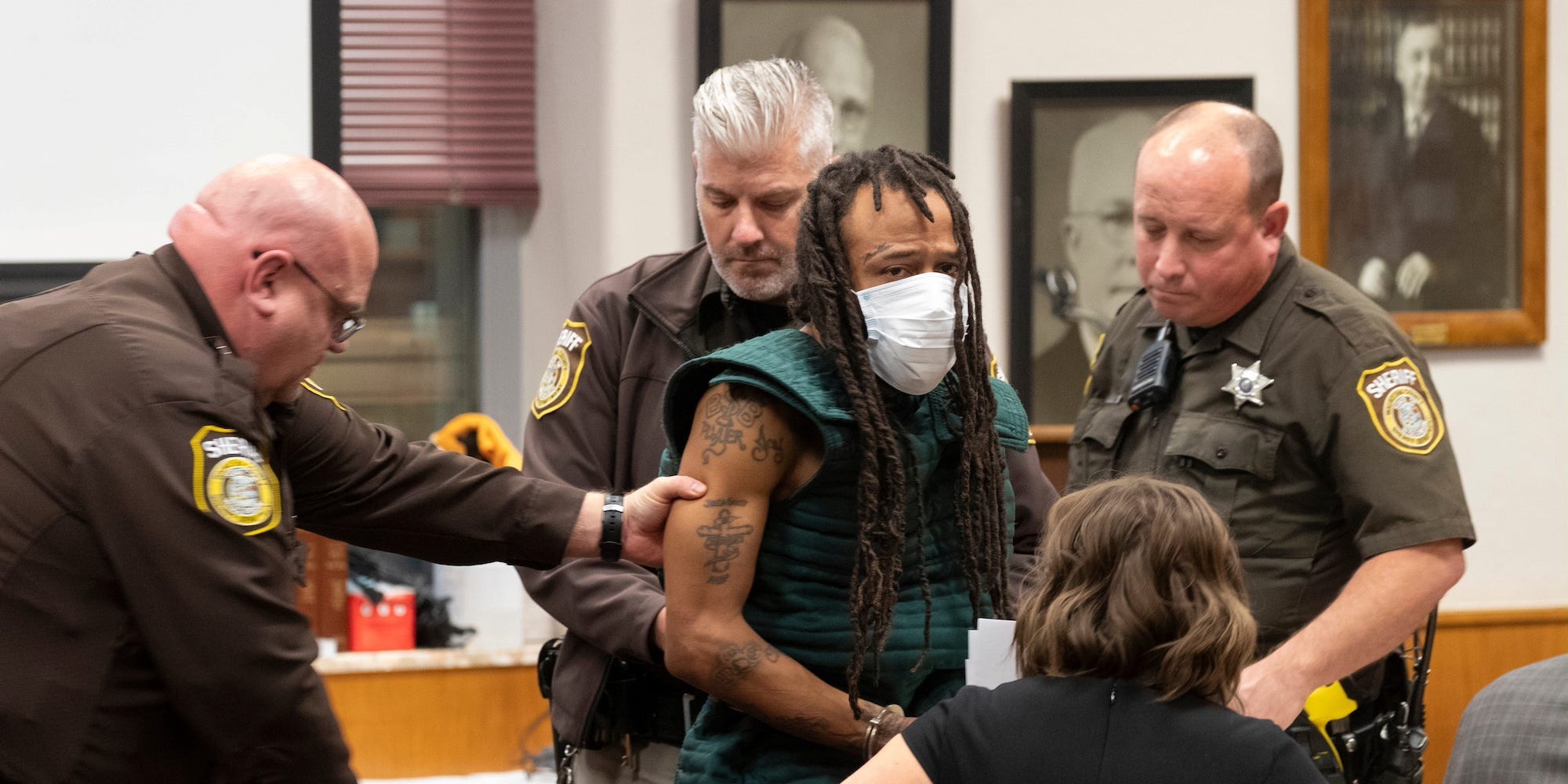 Darrell Brooks, in a mask, joins a court hearing. He is escorted by three officers.